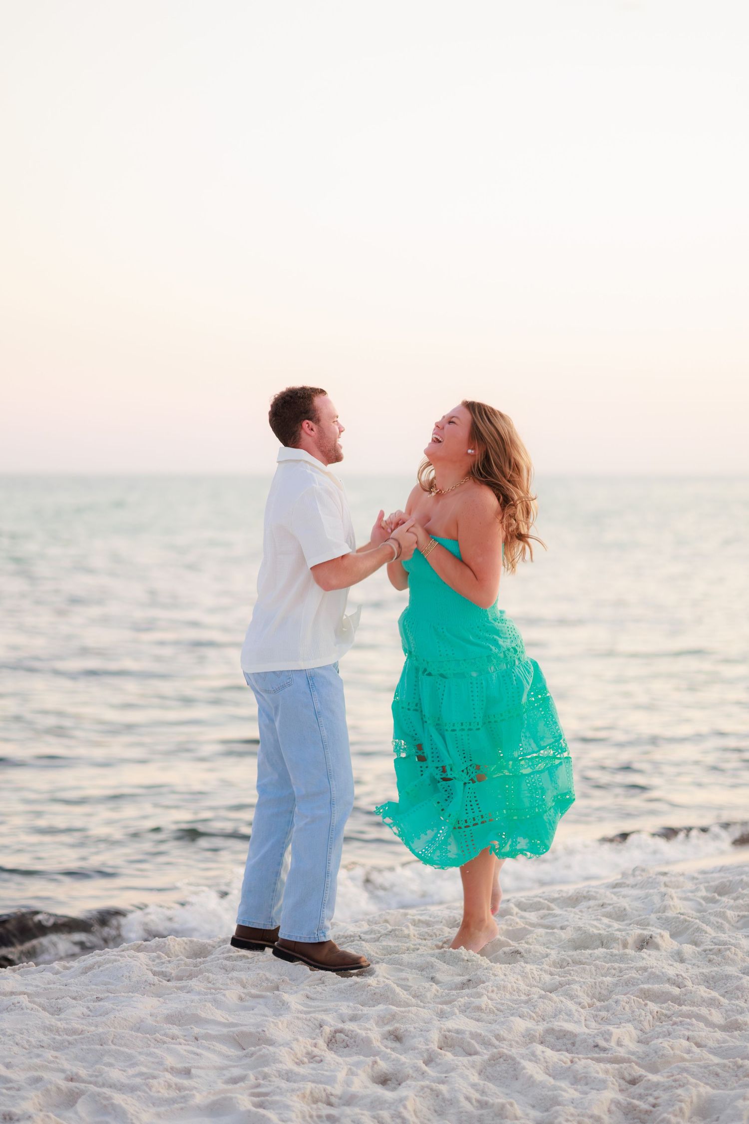 A couple in white and turquoise clothing dance playfully together on a sandy beach at sunset.