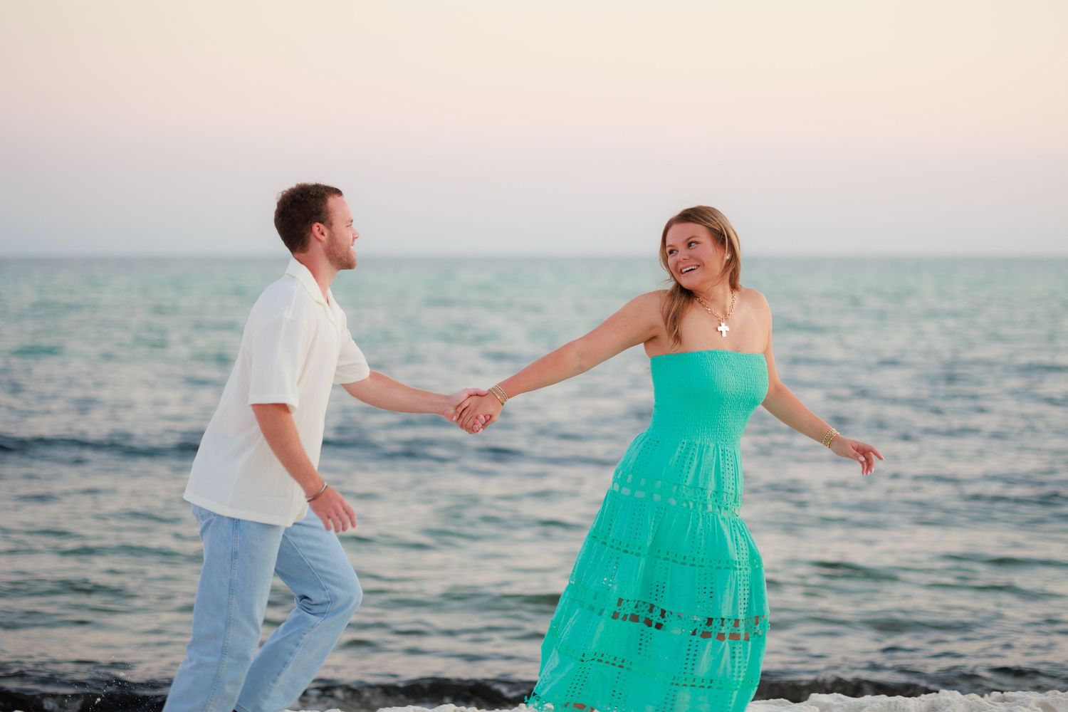 Two people in white and turquoise outfits dance joyfully along the shoreline at dusk.
