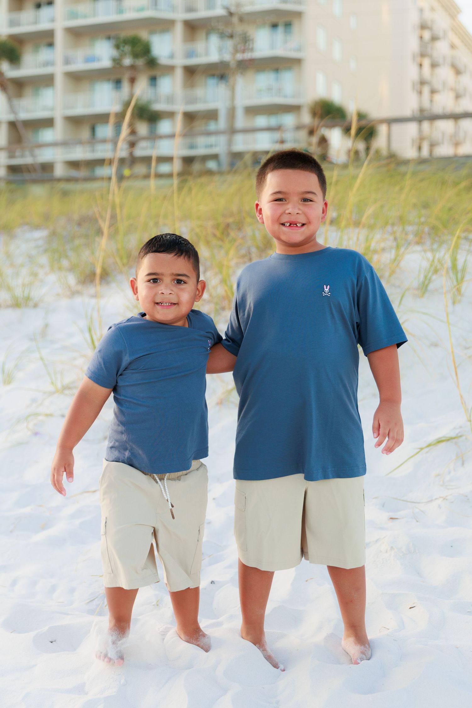 Two children wearing blue shirts and khaki shorts stand together on a white sandy beach with condos in the background.