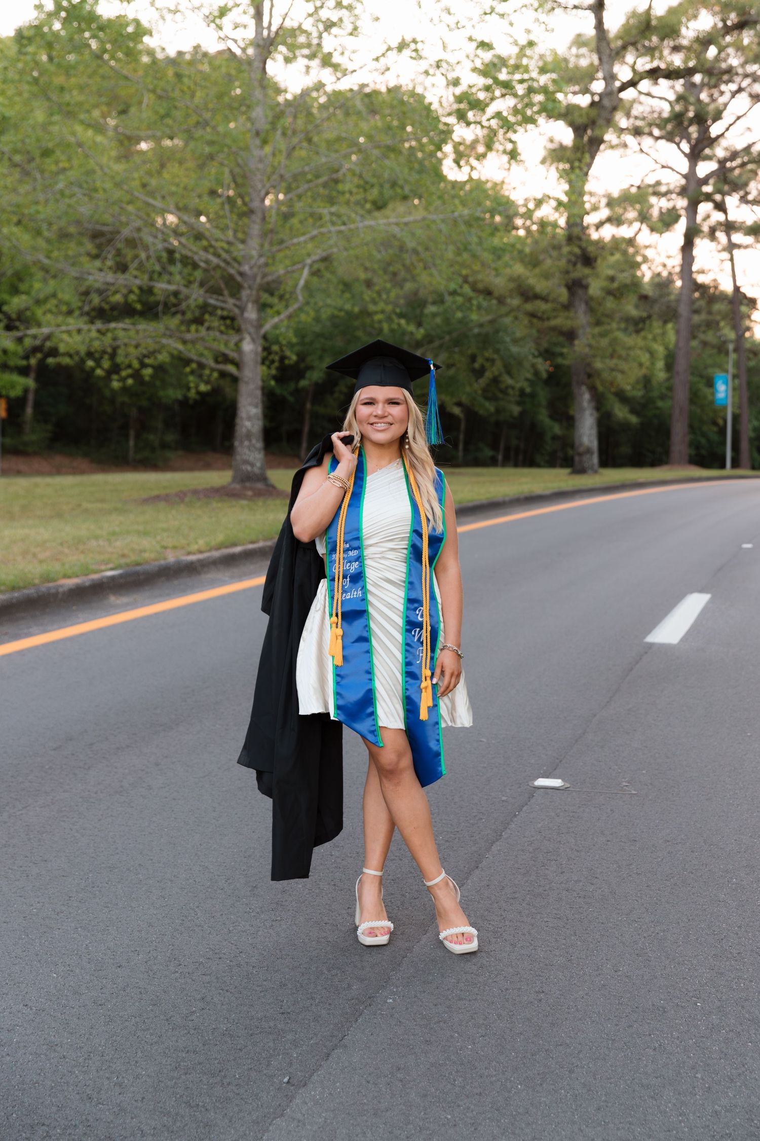 Graduate in cap and gown walks along a tree-lined road during golden hour.