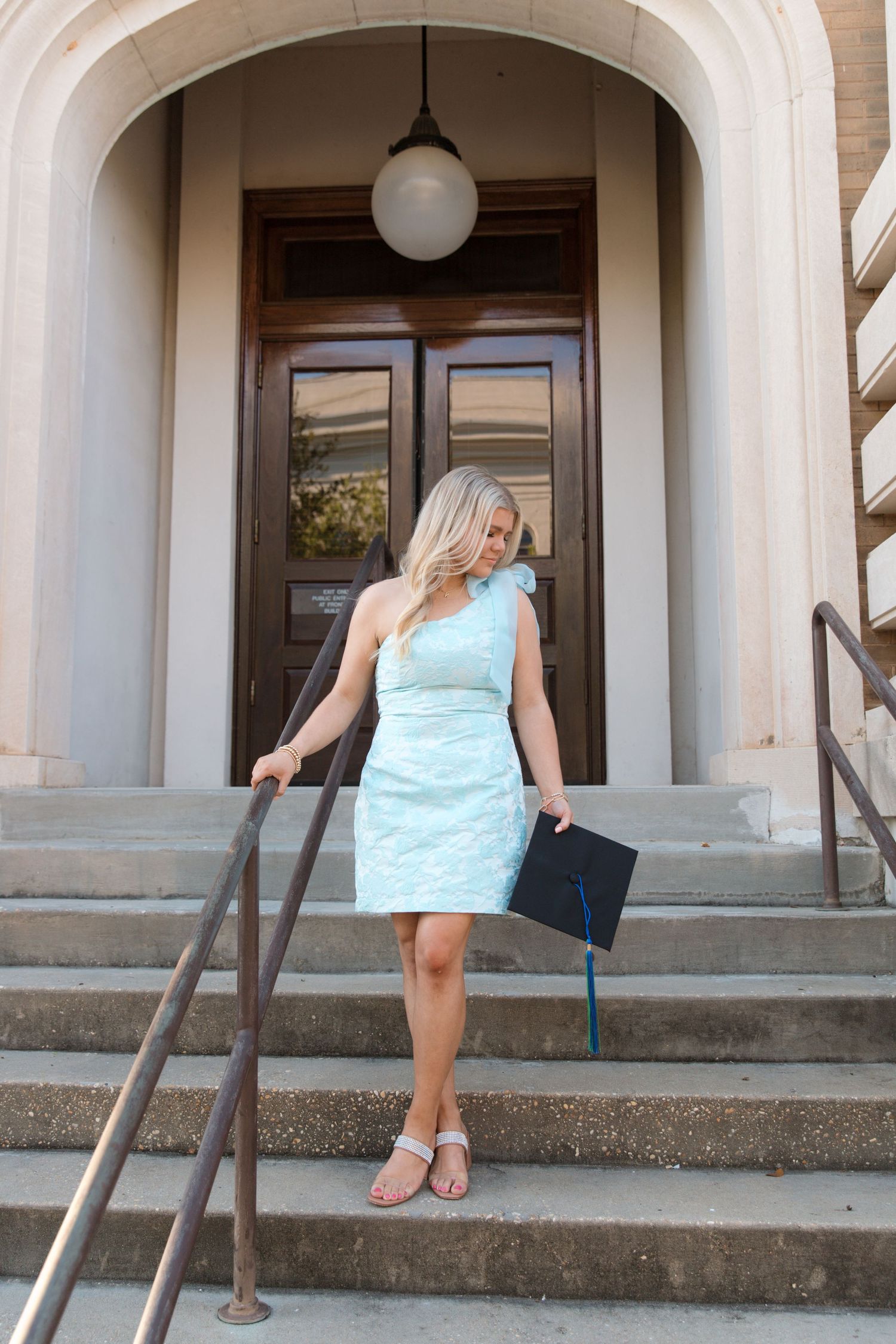 A figure in a light blue dress stands on stone steps in front of a grand wooden door with a globe light fixture above.