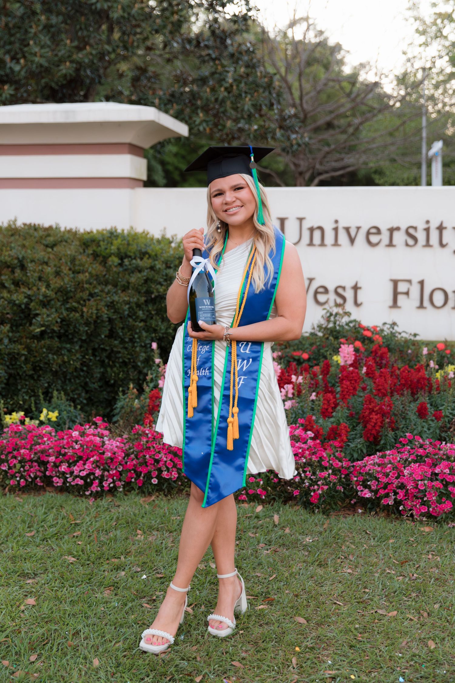 Graduate in blue and white regalia poses for photos in front of vibrant pink flowers during commencement ceremony.