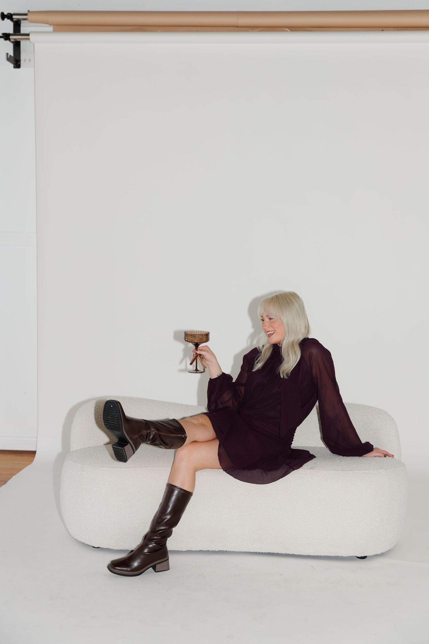 Person in dark dress and boots posing with drink on white modern couch in studio setting.
