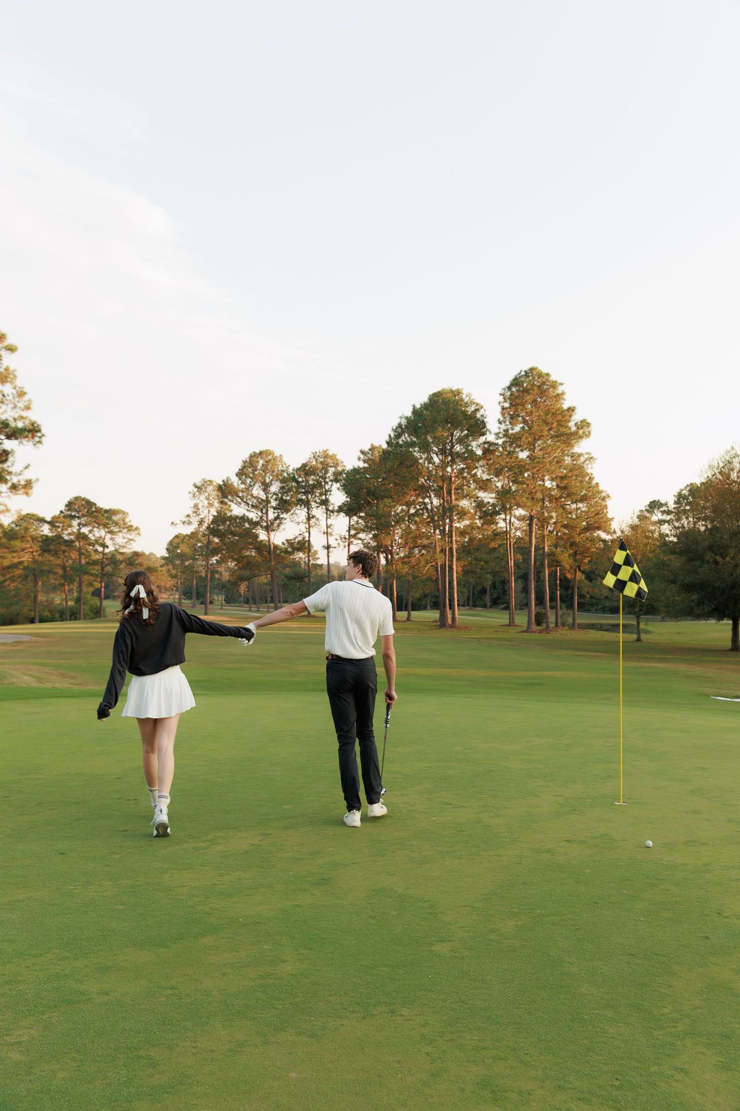 Two people walking on a lush green golf course putting green with a yellow flag pin during golden hour.