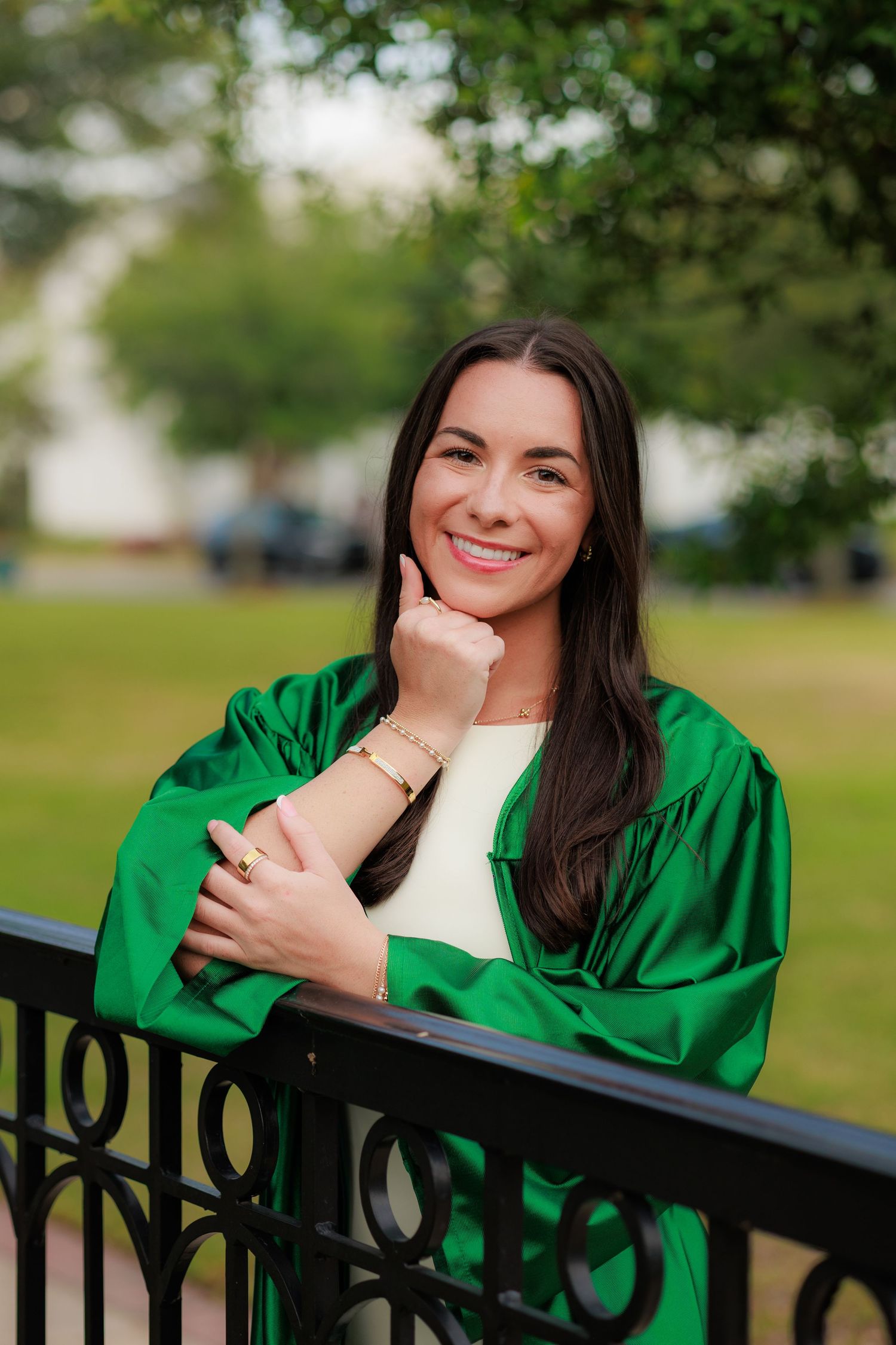 Smiling young woman in green graduation gown posing outdoors by iron fence with lush green trees in background.