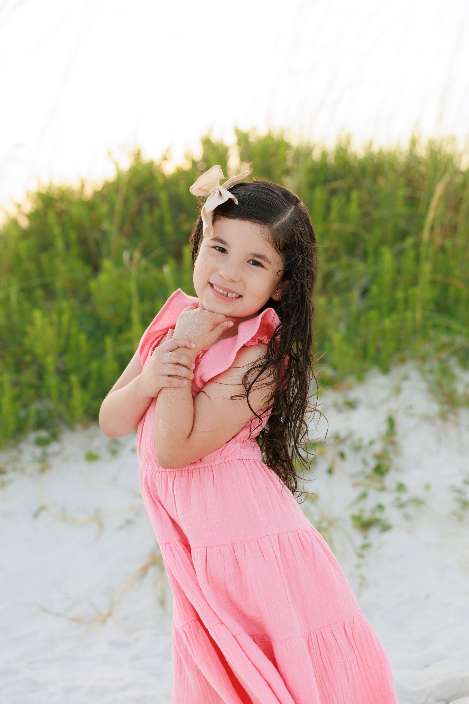 A smiling young girl in a pink dress with a bow in her dark hair poses on white sand dunes with green grass behind her.