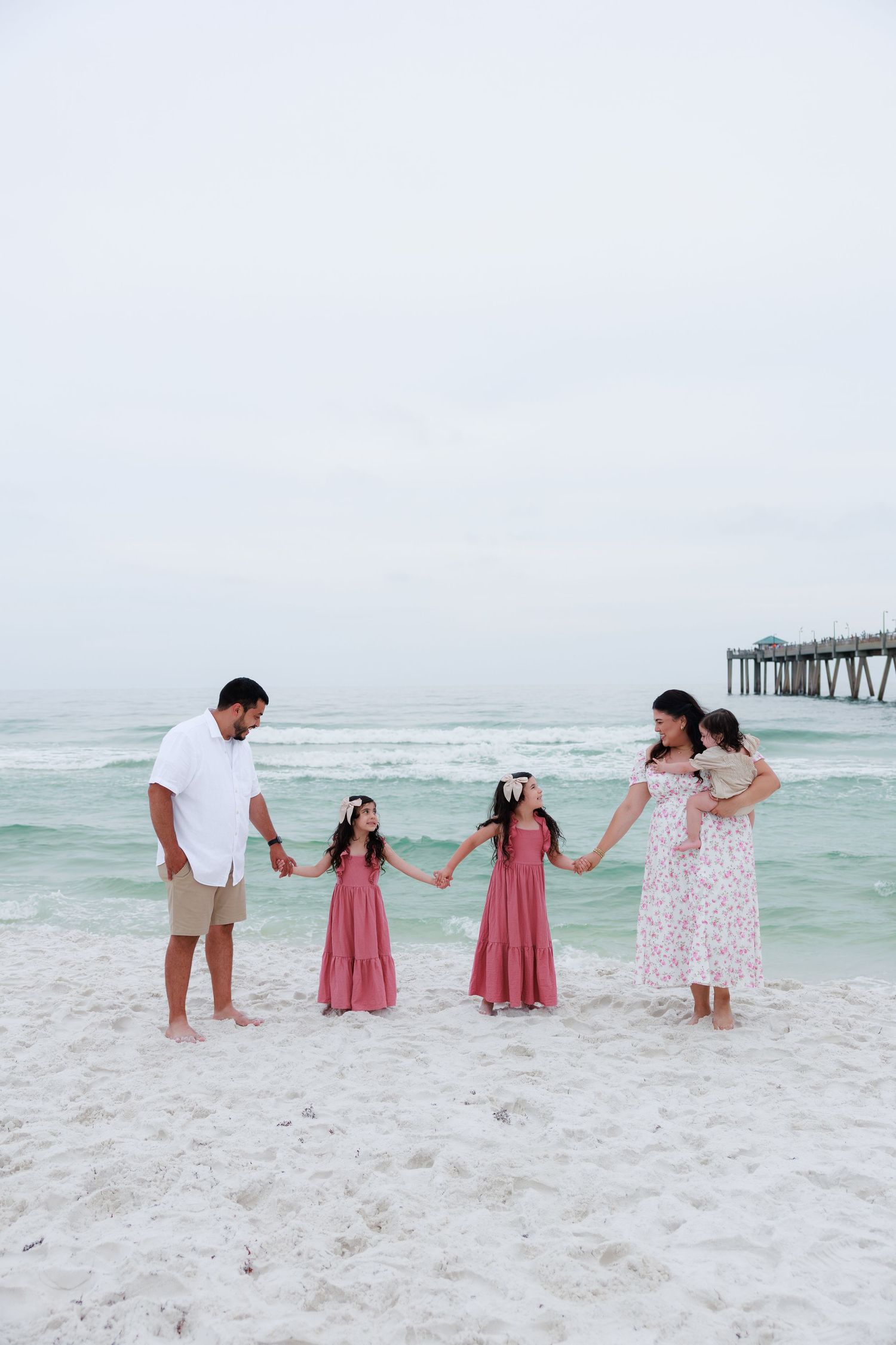 A family of five holds hands on a white sandy beach, three girls in pink dresses with parents near a pier.