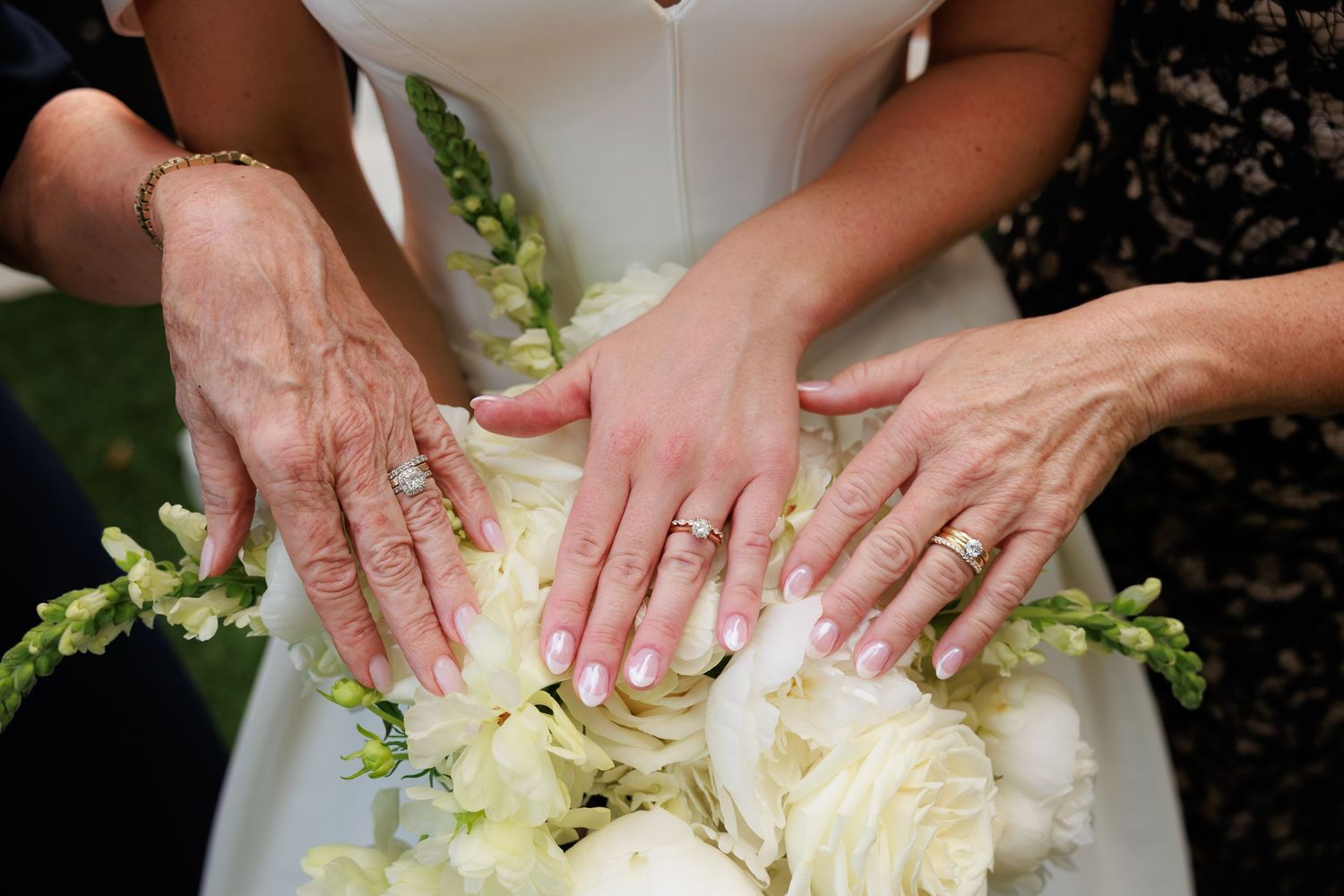 Three generations of women display wedding rings over a white floral bouquet at a wedding ceremony.