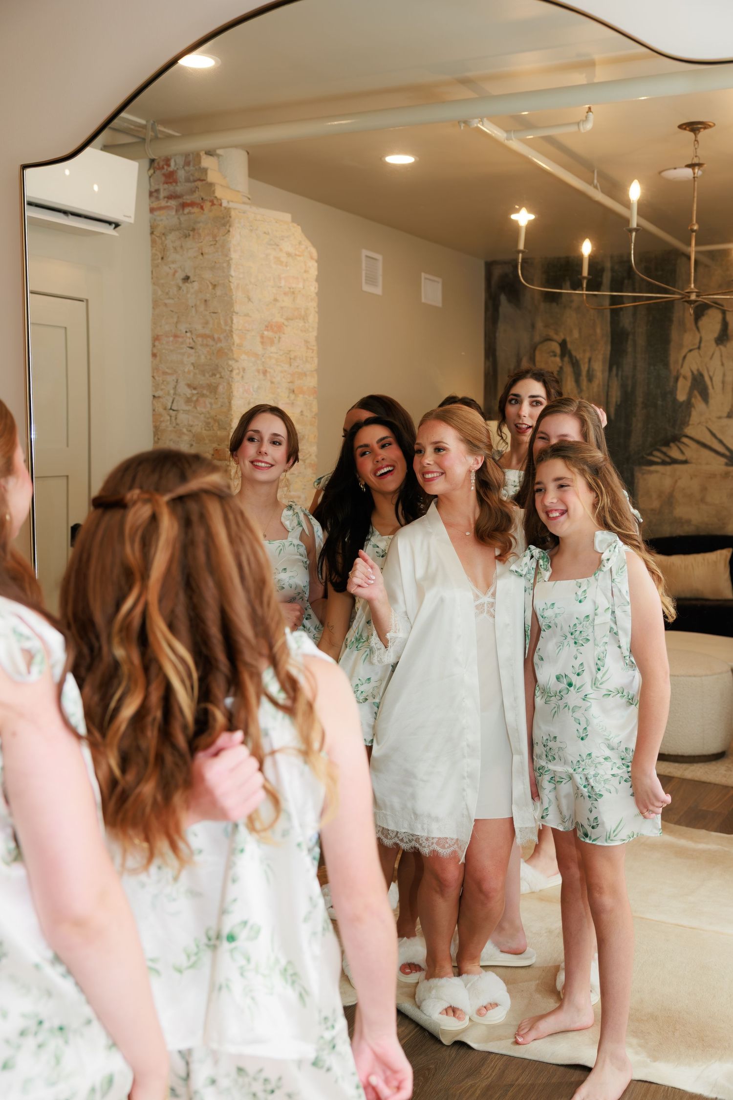 Bridal party in matching floral robes laughing together in mirror reflection during wedding morning preparations.