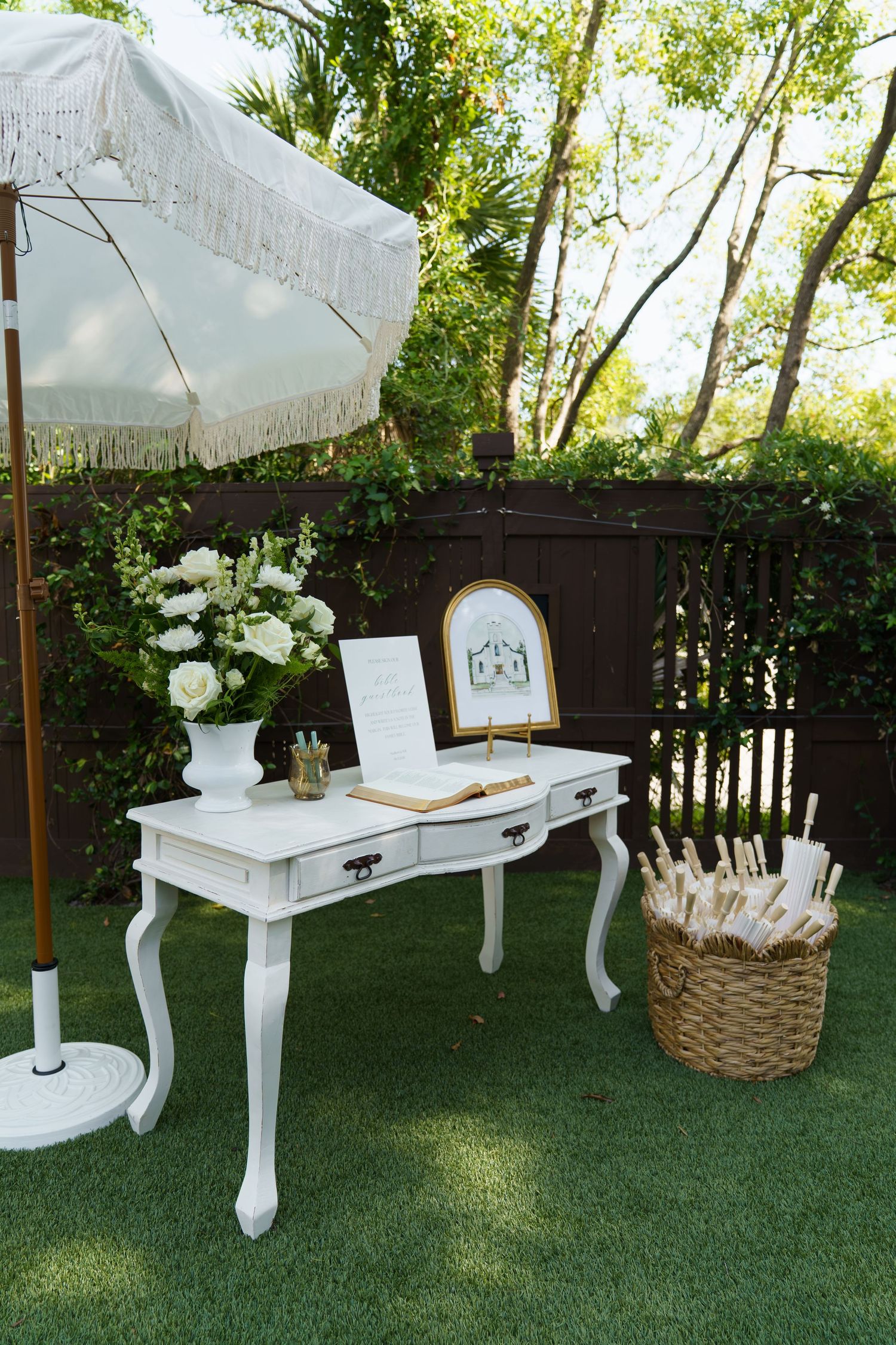 Elegant white vintage desk with guest book, floral arrangement, and wicker basket outdoors under a fringed umbrella.
