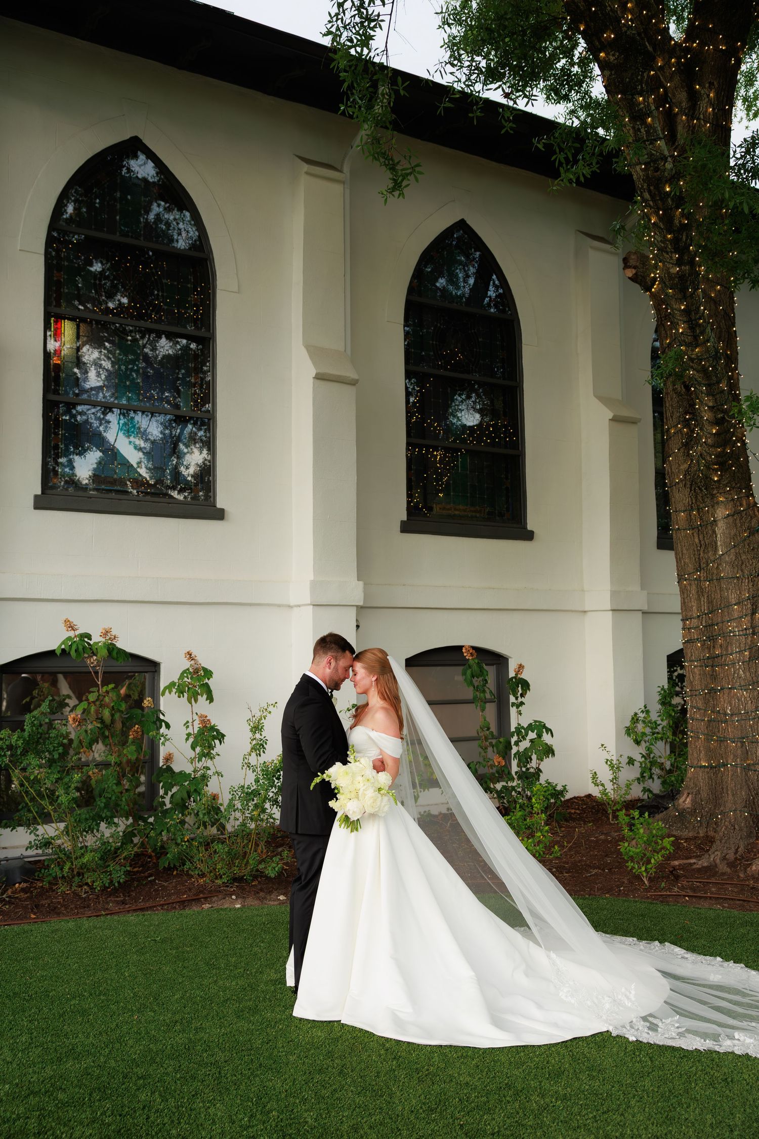 Bride and groom share romantic moment outside white church venue, bride in flowing gown with long veil holding white bouquet.