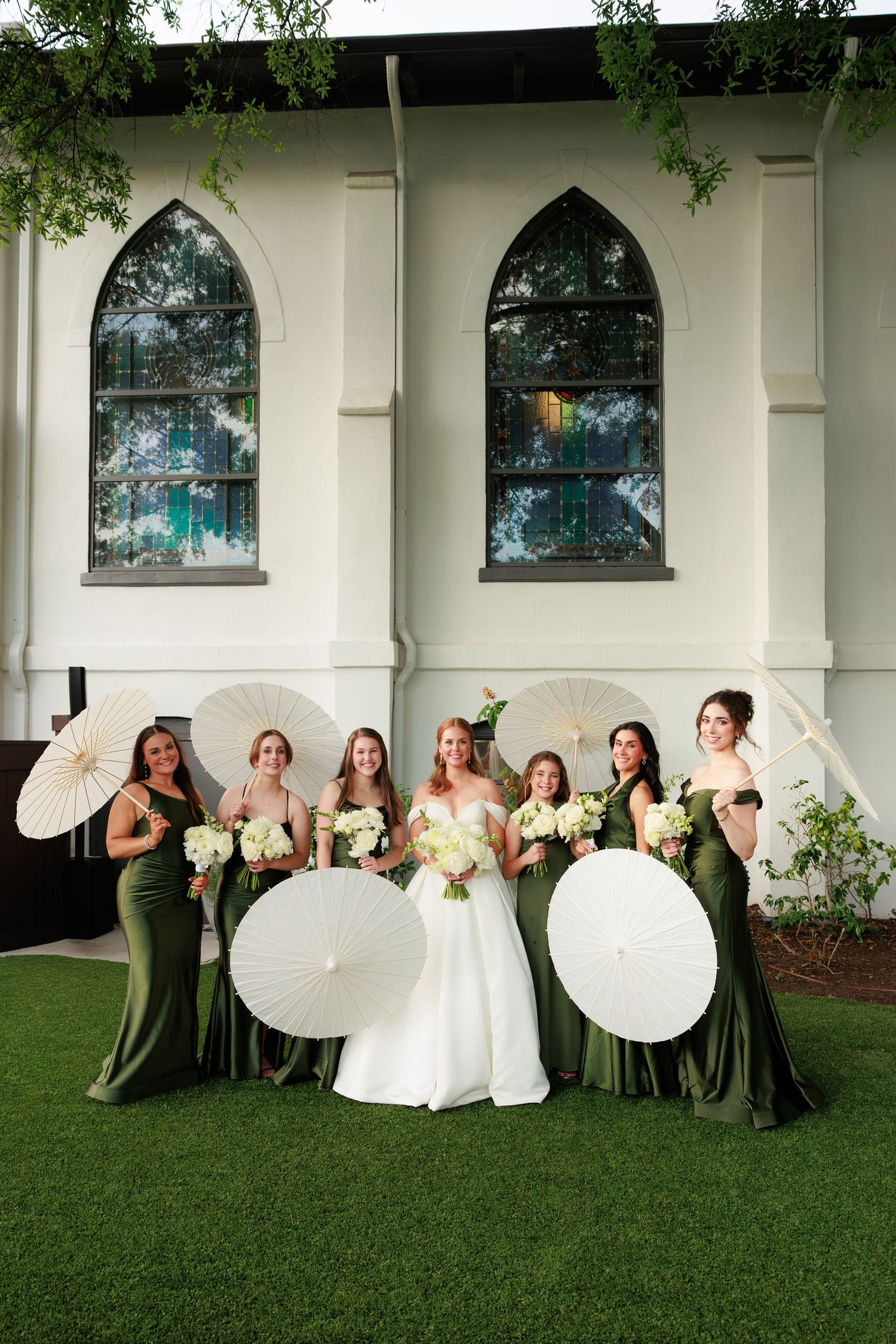 Bride with bridesmaids in olive green gowns holding white parasols and bouquets outside a white chapel.
