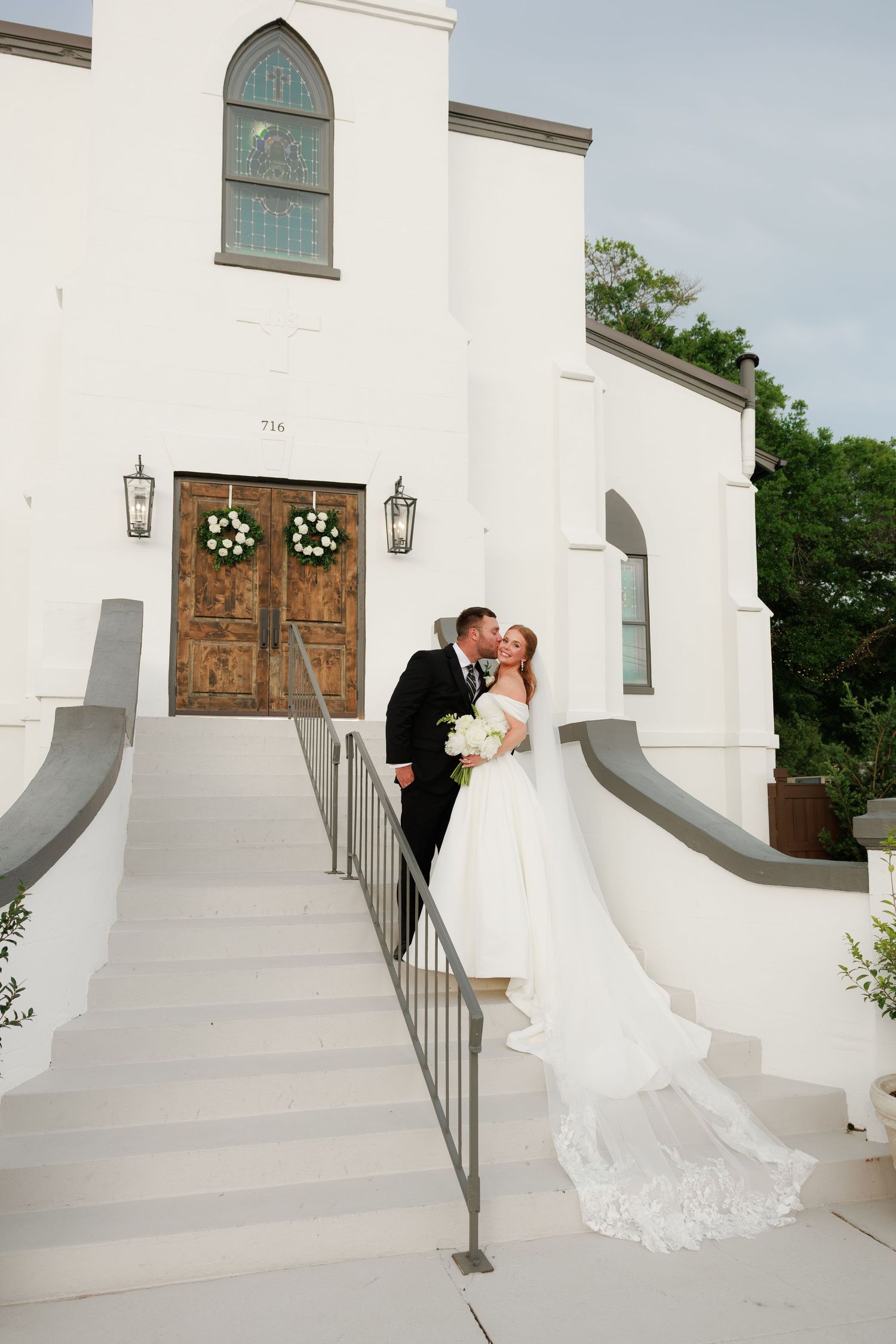 Bride and groom kiss on grand white staircase adorned with wreaths at elegant church wedding venue.