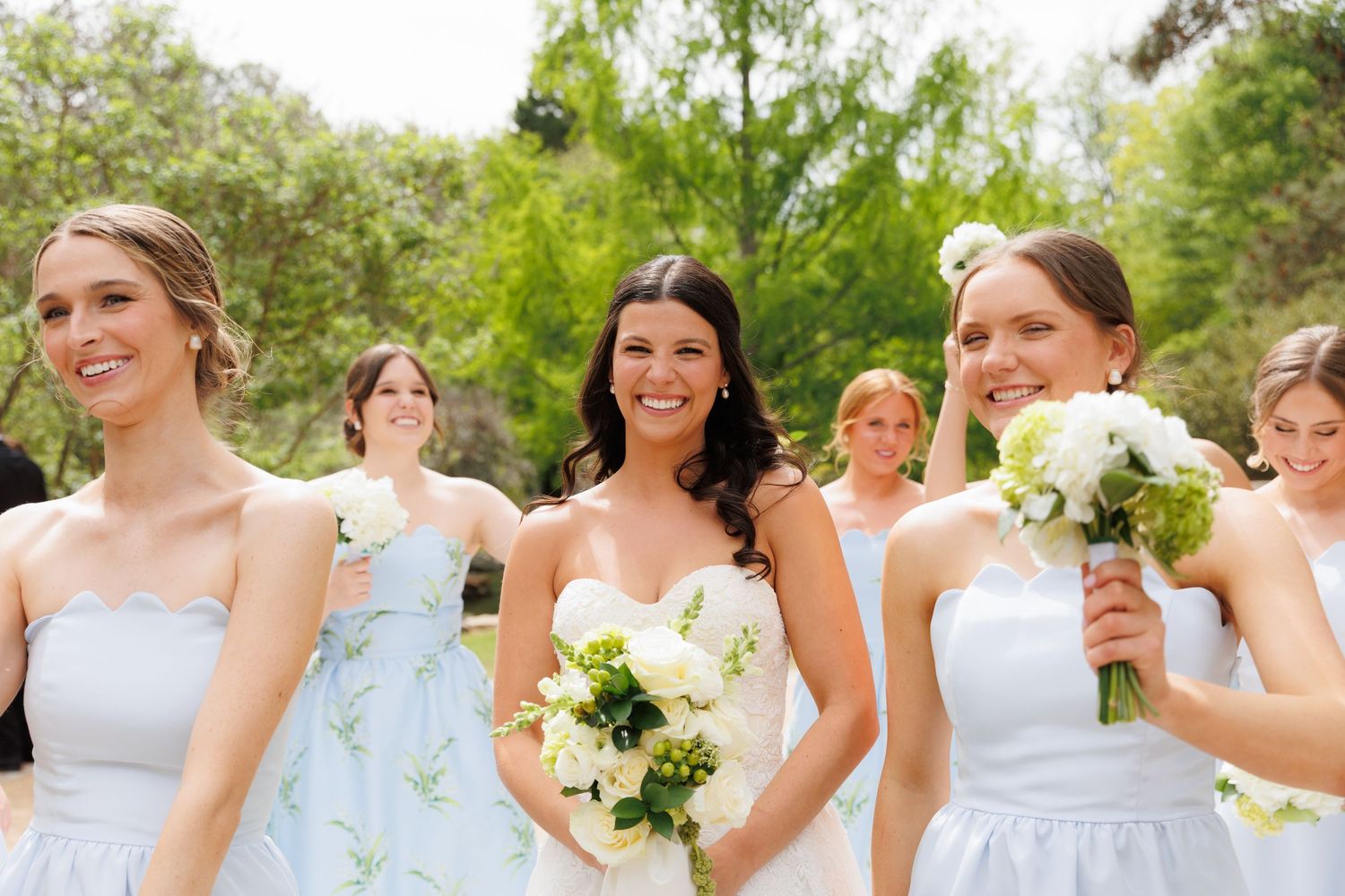 Bride and bridesmaids in light blue dresses laugh together while holding white floral bouquets outside.