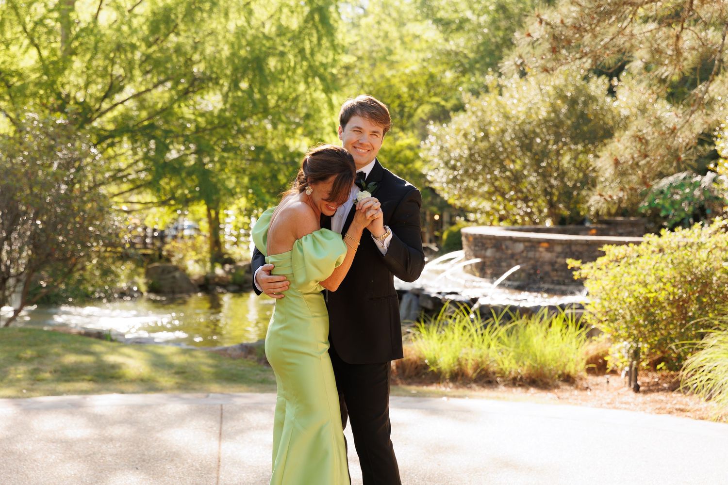 A young couple in formal attire dance together outdoors in a sunny garden setting with lush trees.