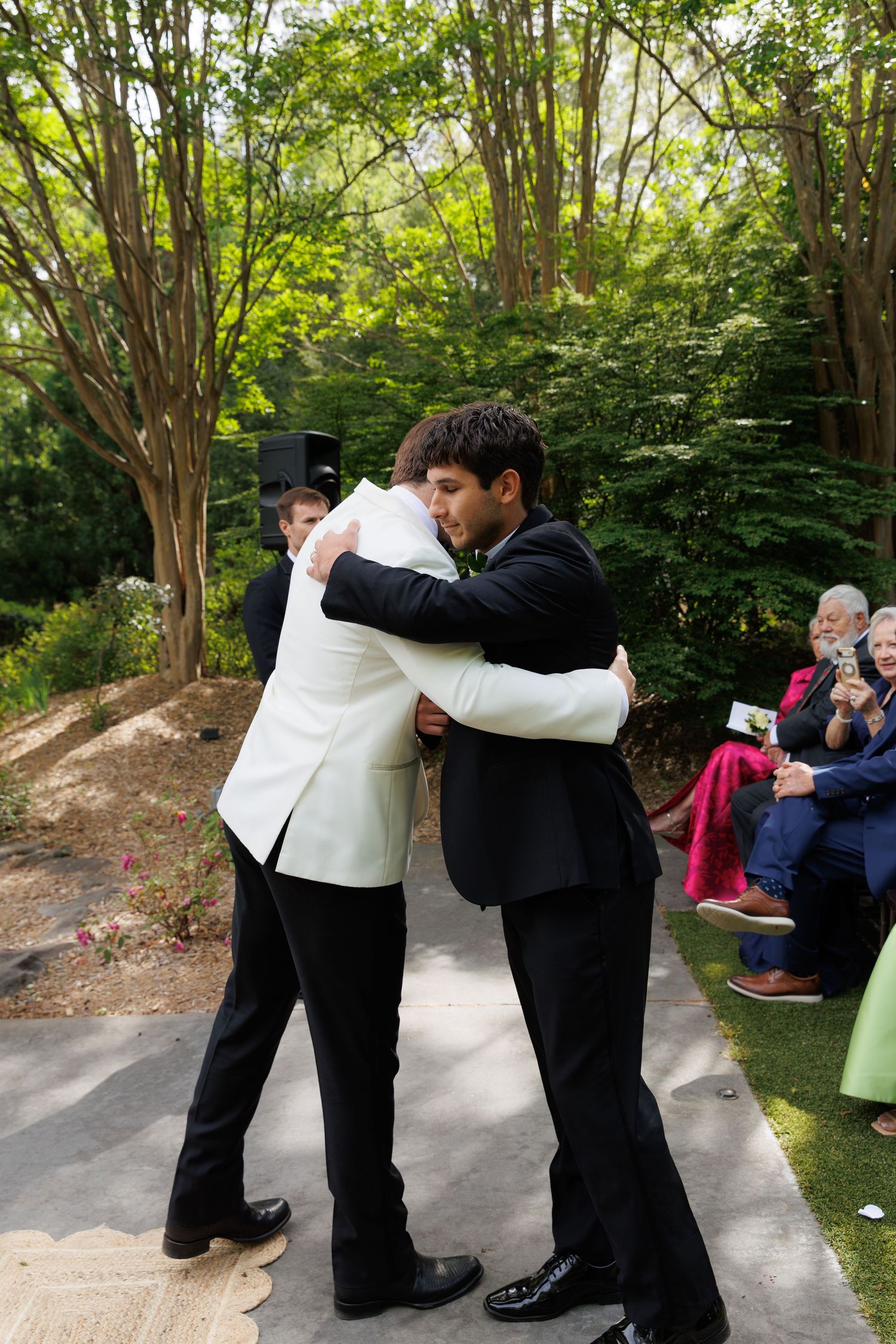 Two grooms share an emotional hug during their outdoor wedding ceremony surrounded by lush green trees.