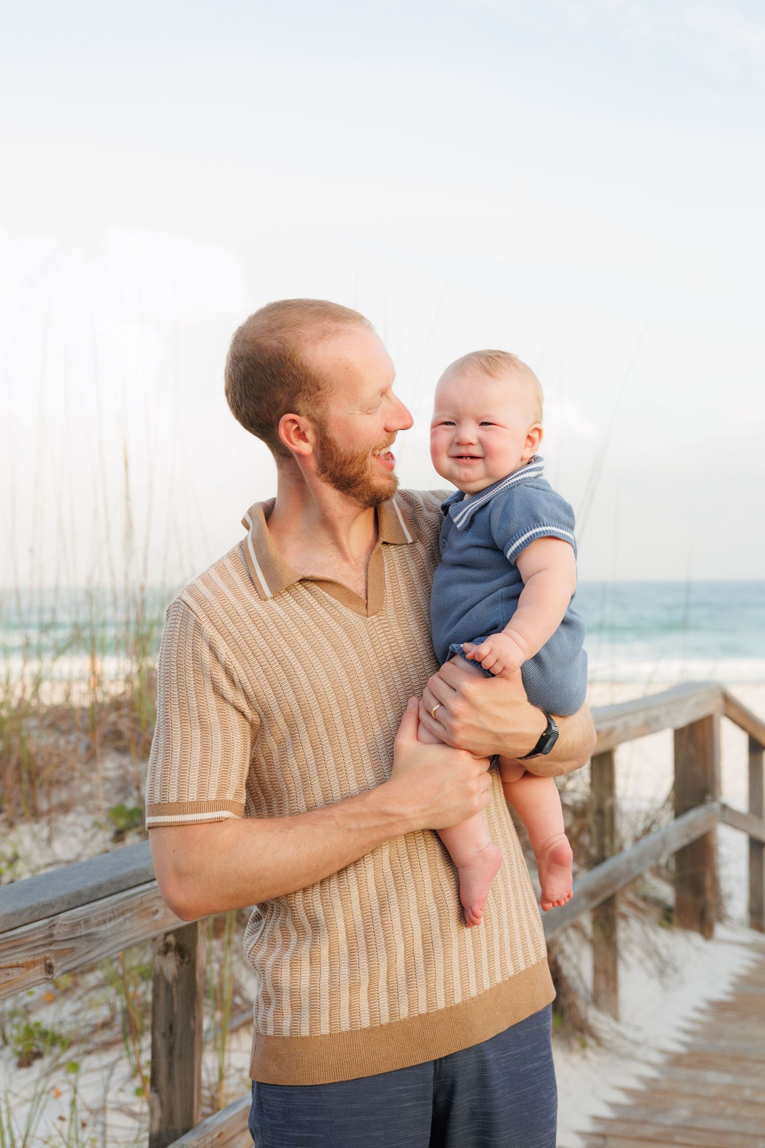 Father holding smiling baby boy on sunny beach boardwalk with ocean and sand dunes behind them.