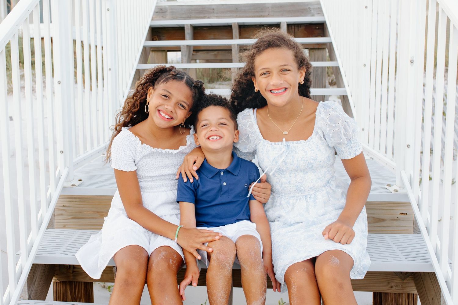 Three siblings in white outfits smiling together on white wooden stairs.