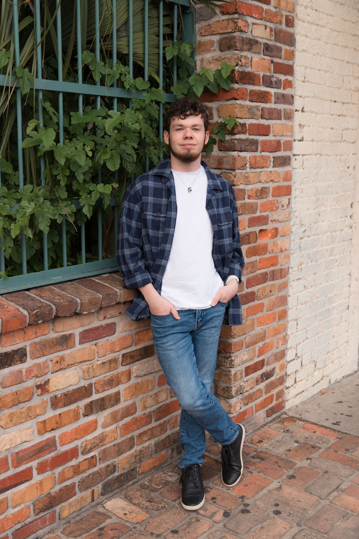 Standing by brick wall and iron fence in casual plaid shirt and jeans.