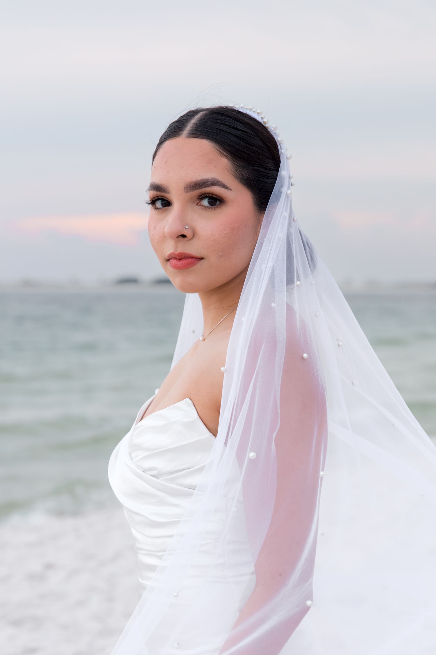 Bride wearing long veil and strapless gown poses against ocean backdrop.