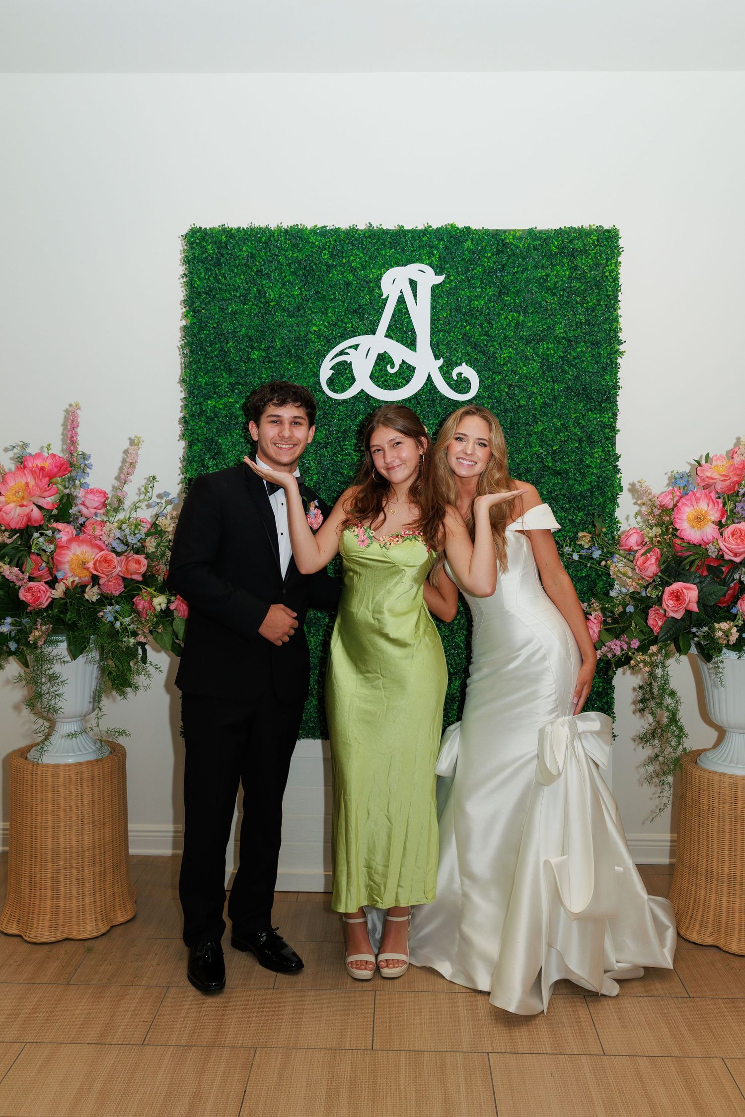 Group poses in formal attire against a green hedge wall with floral arrangements and a decorative monogram letter.