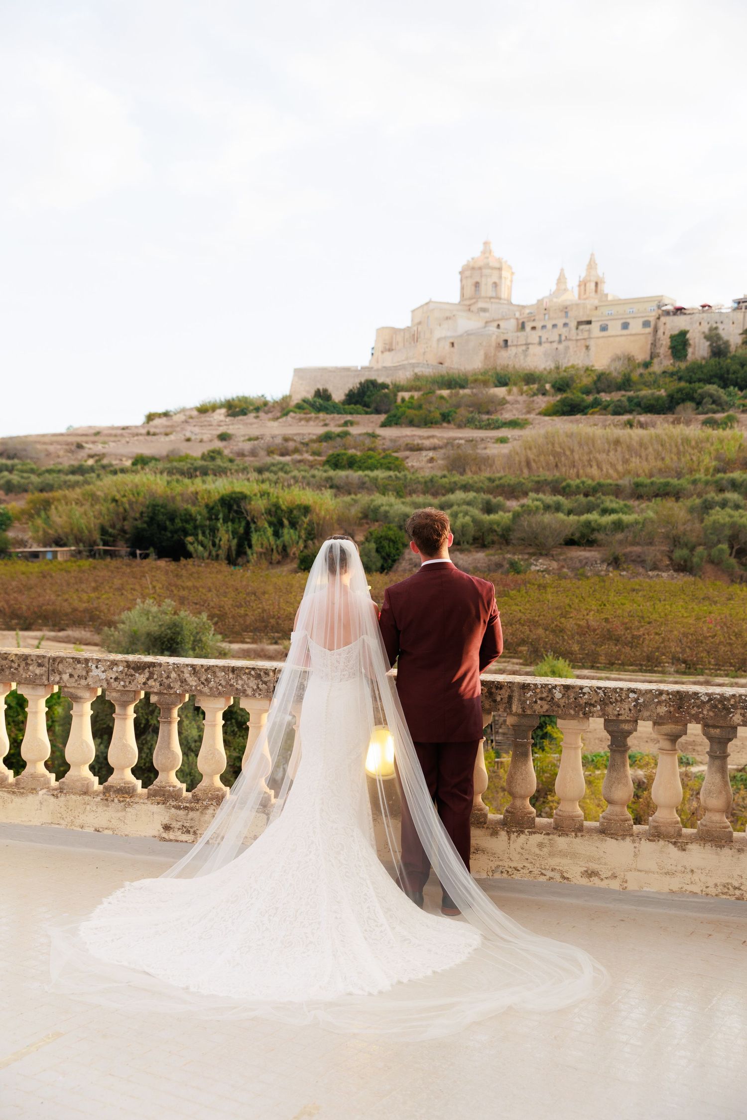 A flowing white wedding veil cascades across a stone balcony with a historic building visible on the hilltop.