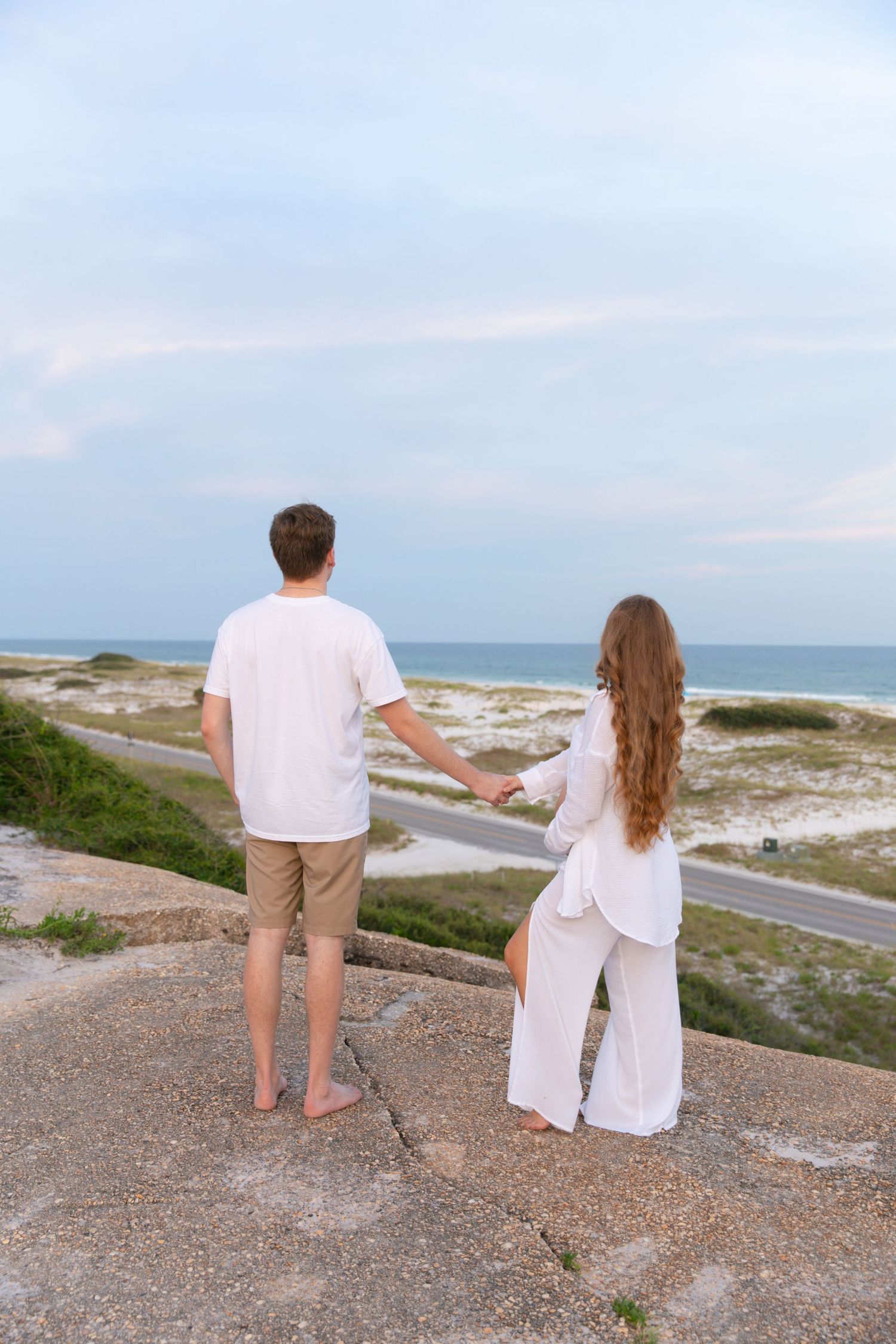 A couple in white attire sits together on a rocky overlook enjoying scenic coastal views at sunset.