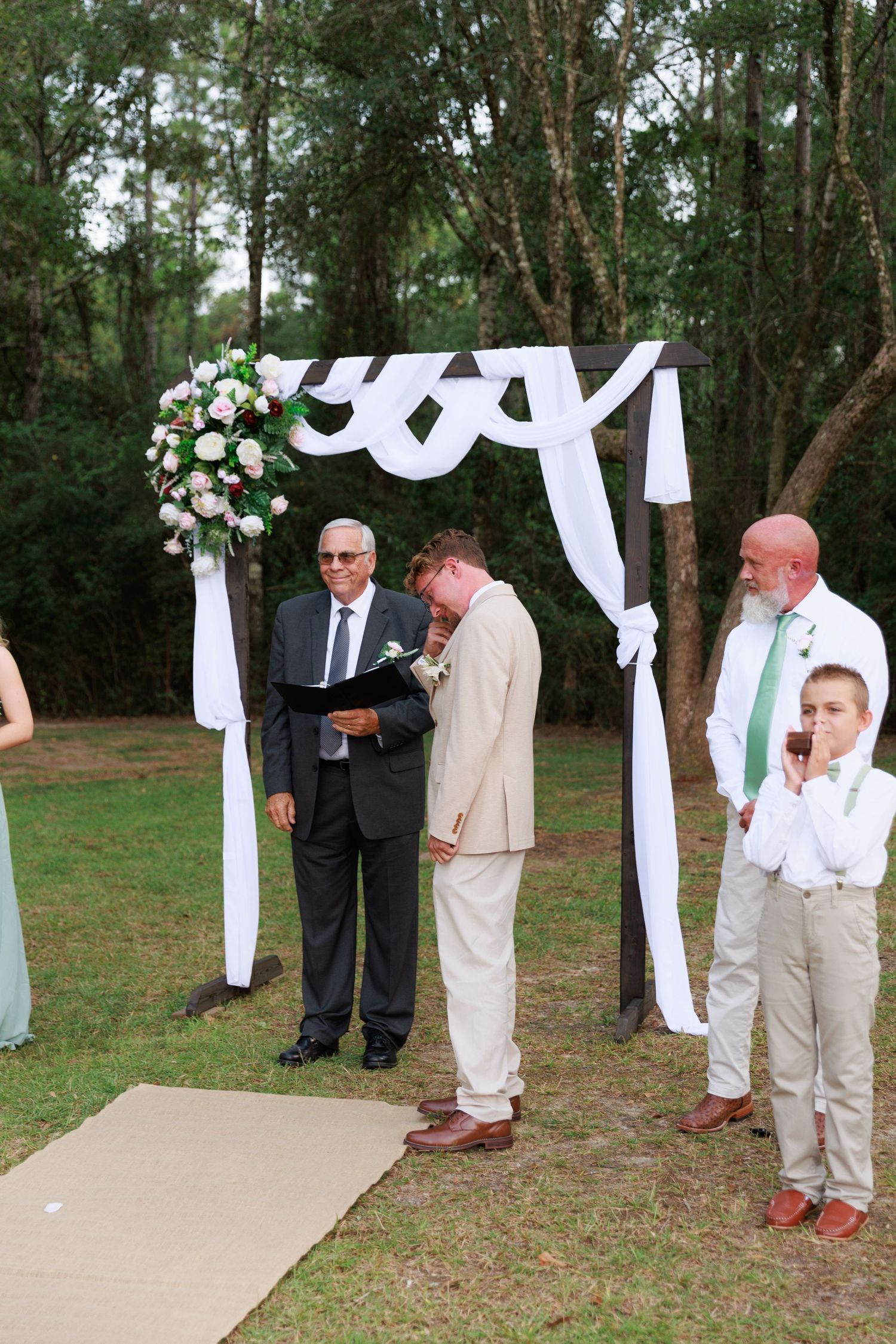 A wedding ceremony in progress under a white fabric arch decorated with pink and white flowers.