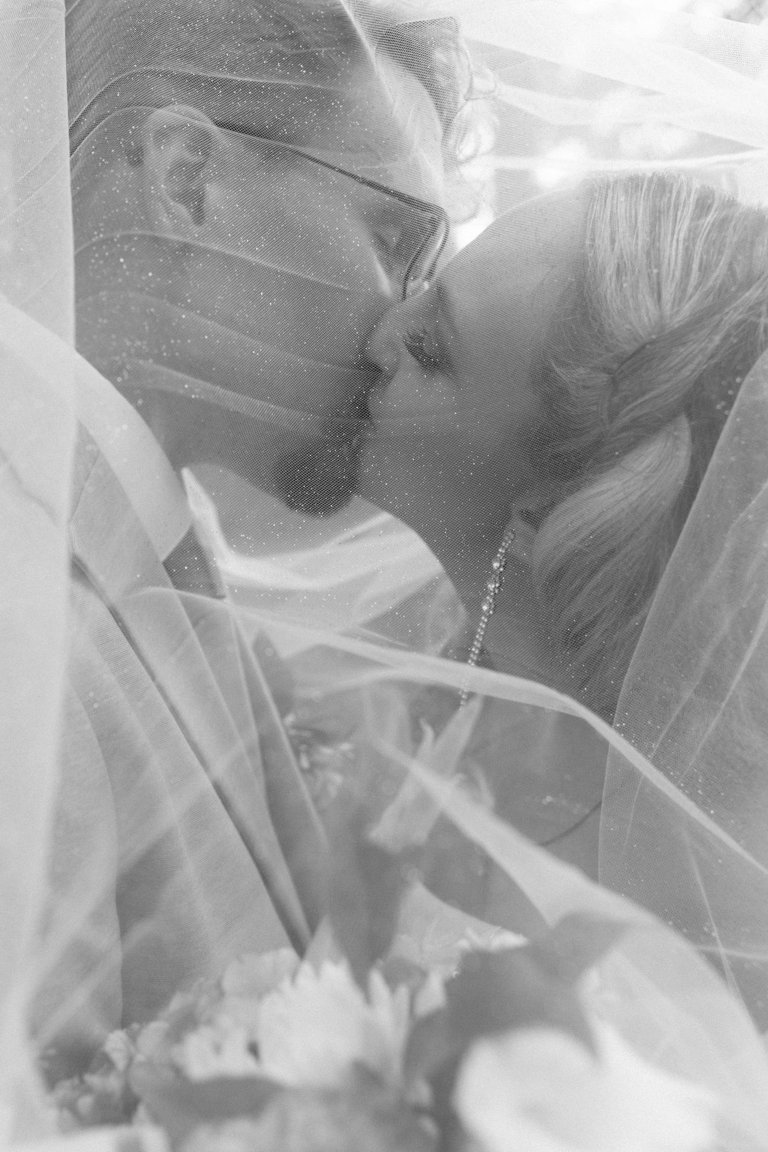 Black and white romantic wedding photo of couple sharing intimate moment under flowing veil.