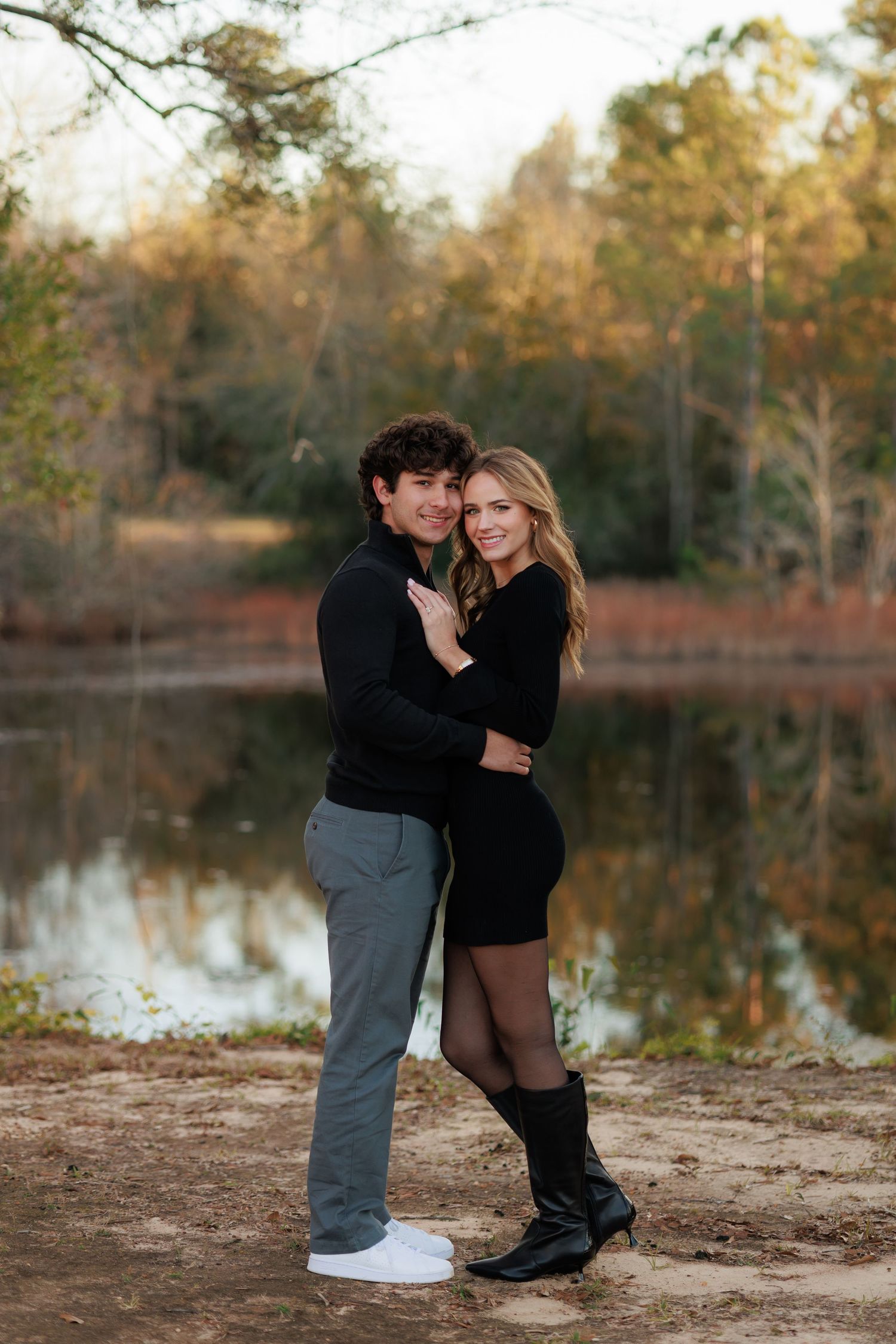 Romantic couple embraces by a serene lake during golden hour with autumn trees reflecting in the calm water.