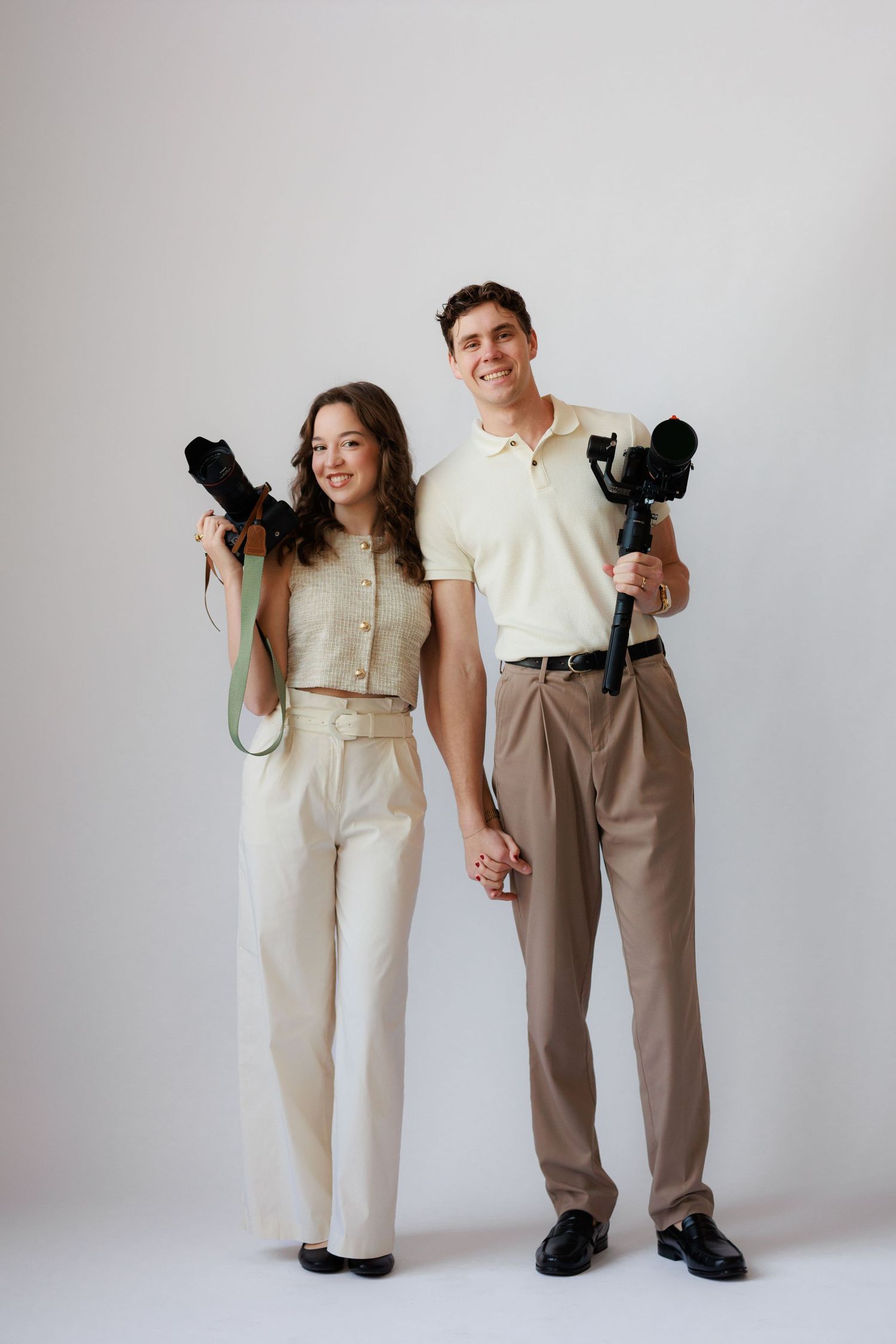 Two people in contrasting beige and white outfits hold black cameras while posing against a light background.