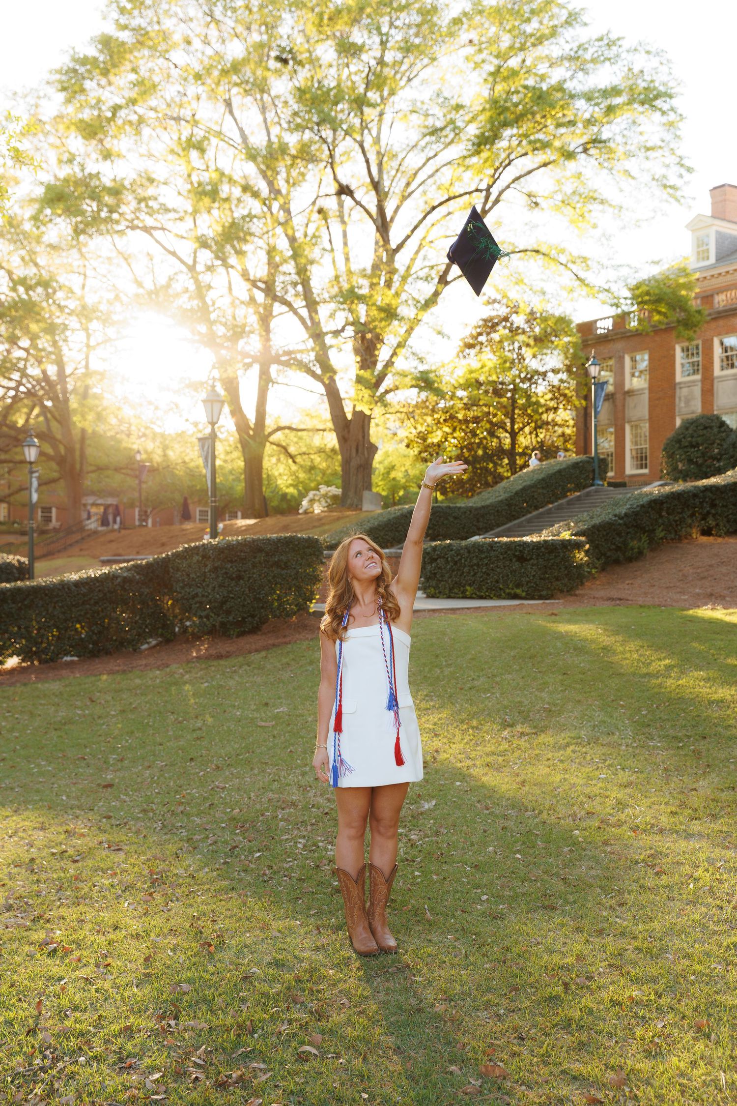 Joyful graduate in white dress tosses mortarboard cap into the air on sunny college campus.