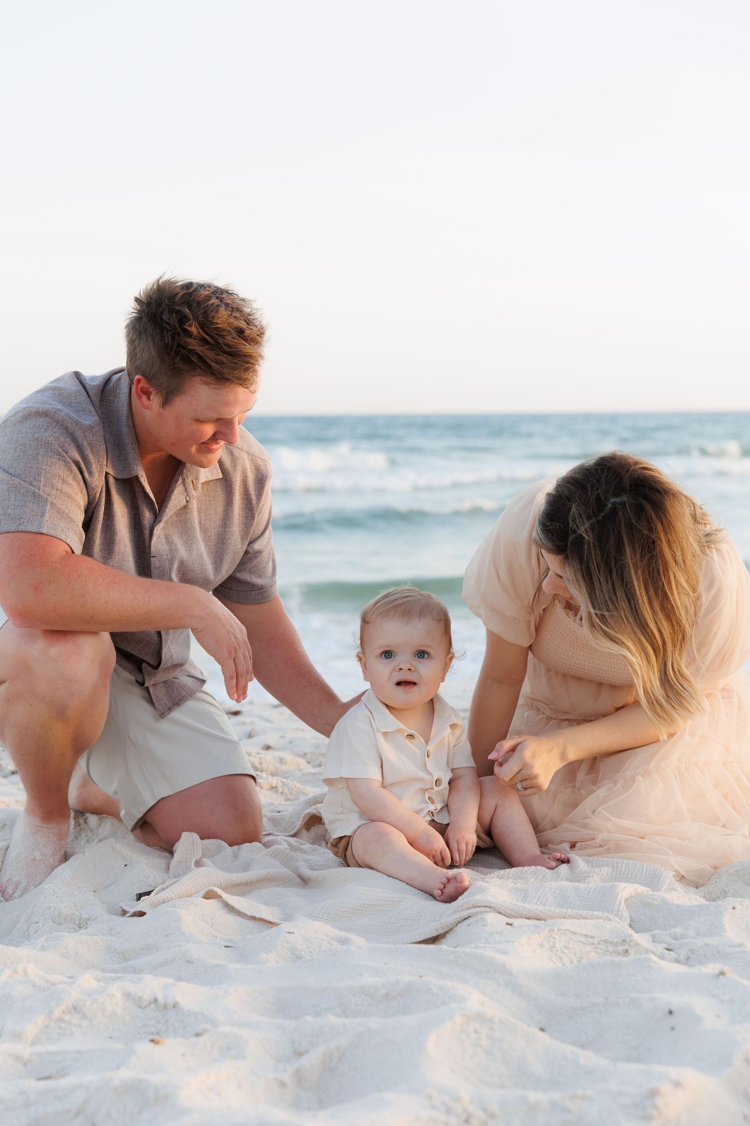 Young family with baby sitting on white sand beach near ocean waves at sunset.