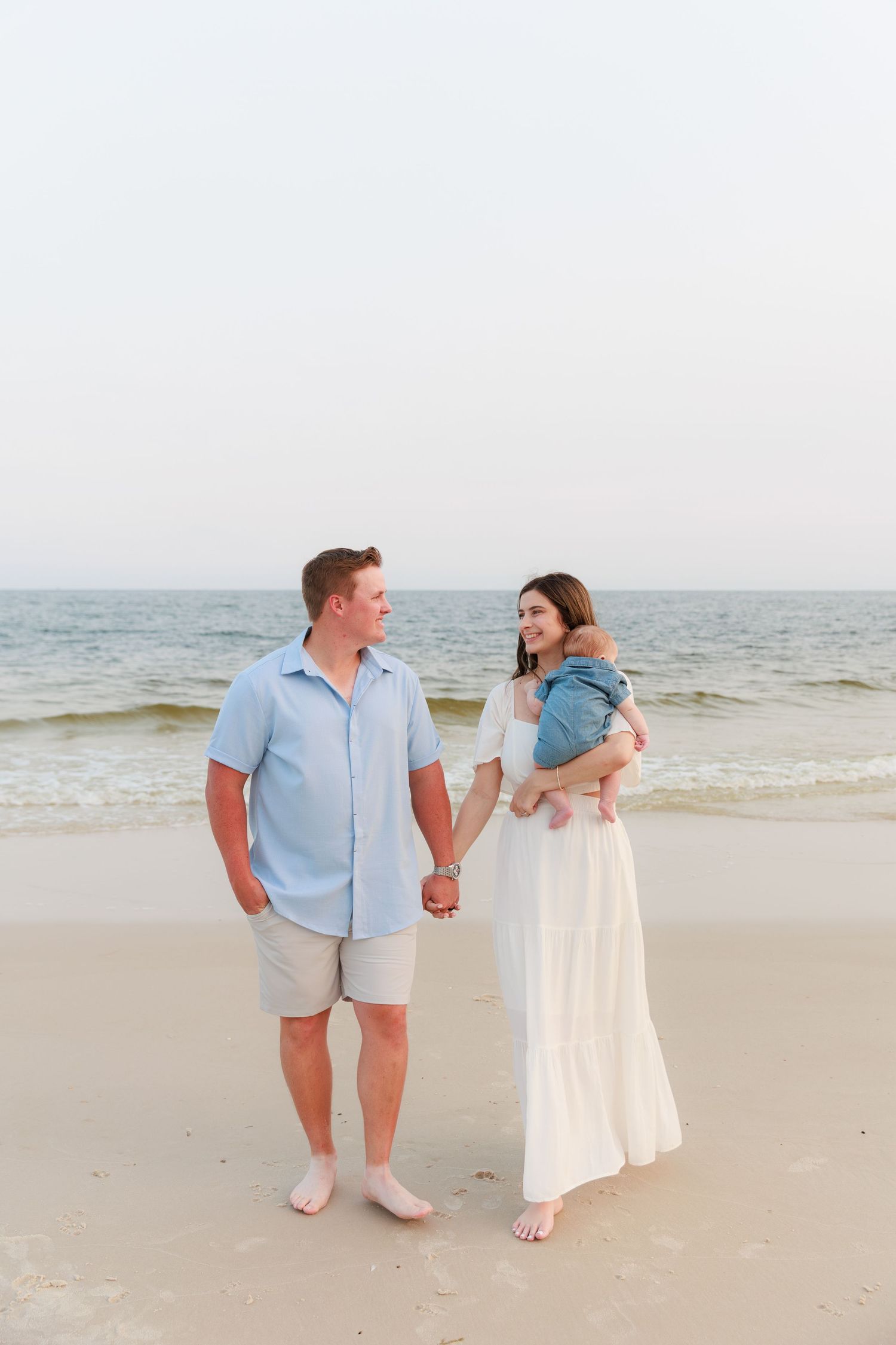 A couple holds hands on a sandy beach, the woman in a white dress holding a baby, the man in a light blue shirt.