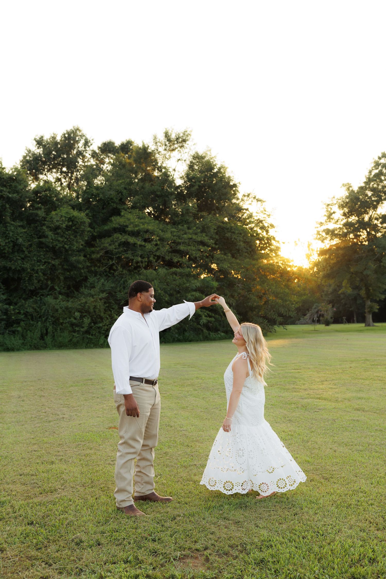 Couple dancing at sunset in a green field, man in white shirt twirling woman in white lace dress.