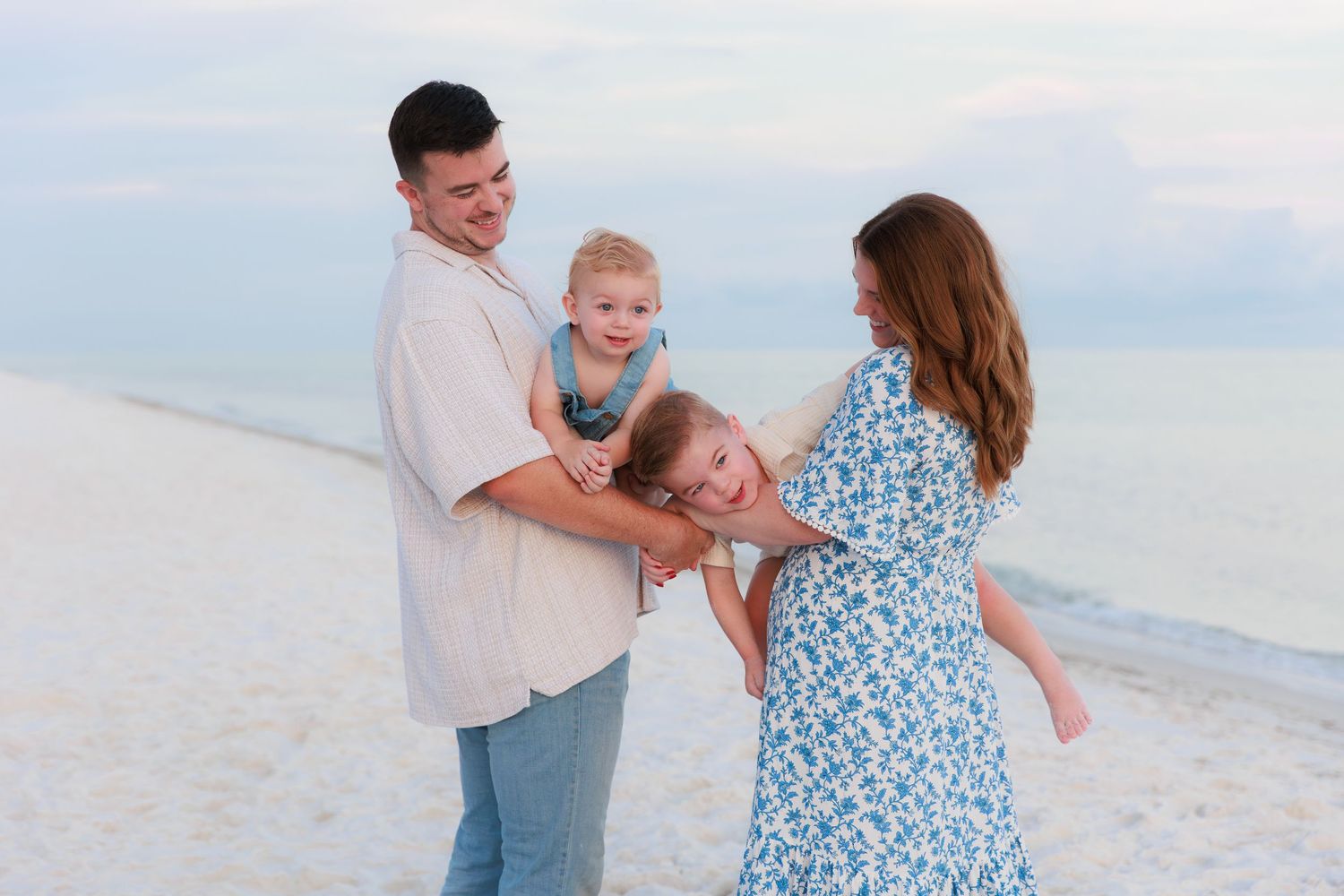 Family of four enjoying a beach day, parents playfully swinging children on white sand shore.