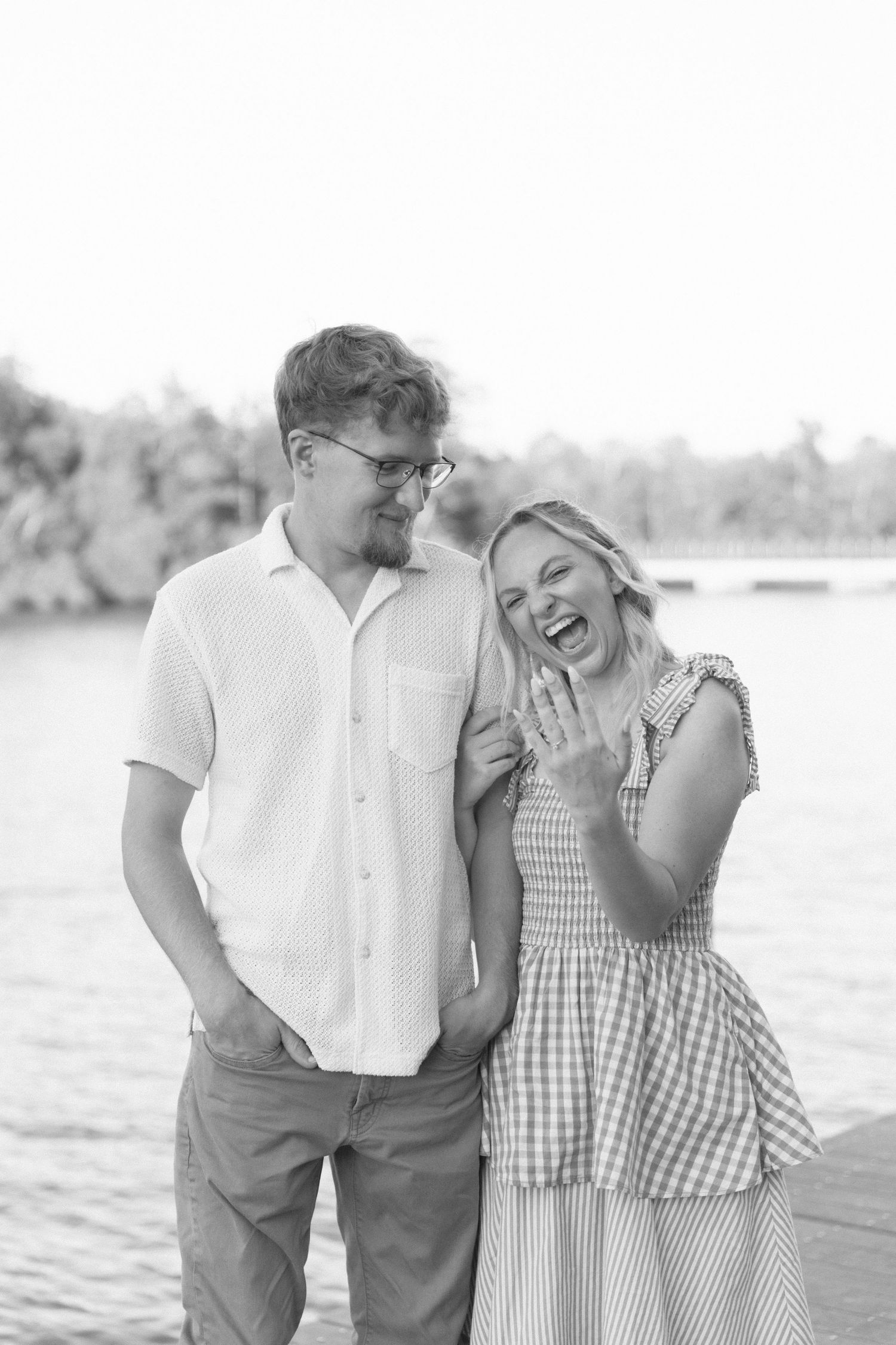 Black and white photo of a laughing couple on a dock, woman showing engagement ring, trees and lake in background.