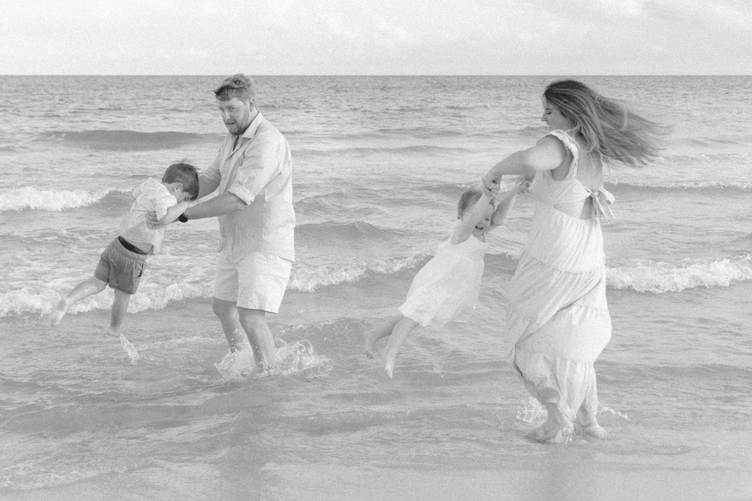 Black and white photo of a joyful family playing in ocean waves on a sandy beach.