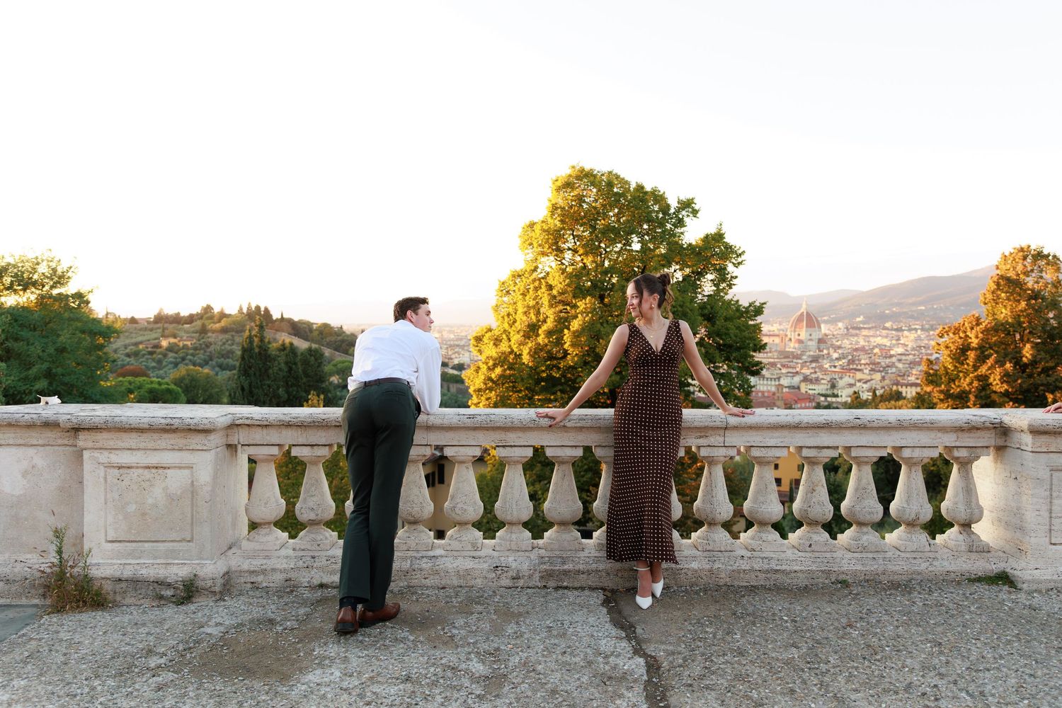 Couple standing at a stone balustrade terrace overlooking Florence cityscape at golden hour sunset.