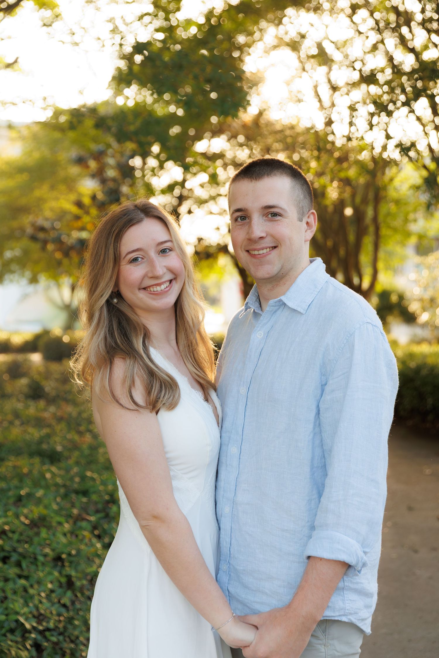 A smiling young couple poses outdoors in golden hour light, woman in white dress and man in light blue shirt.