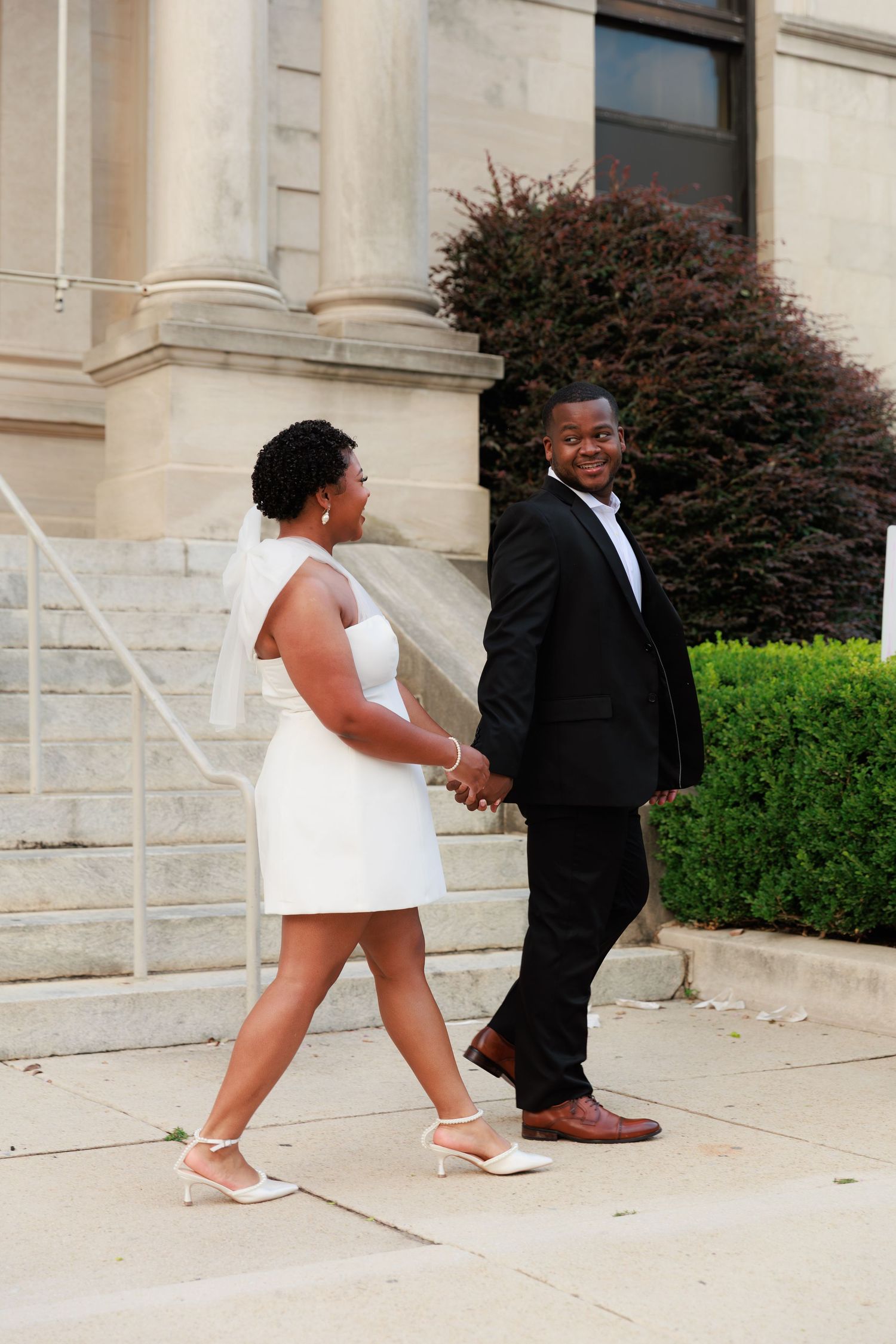 A newlywed couple in wedding attire holds hands and walks outside a stone building, smiling joyfully.