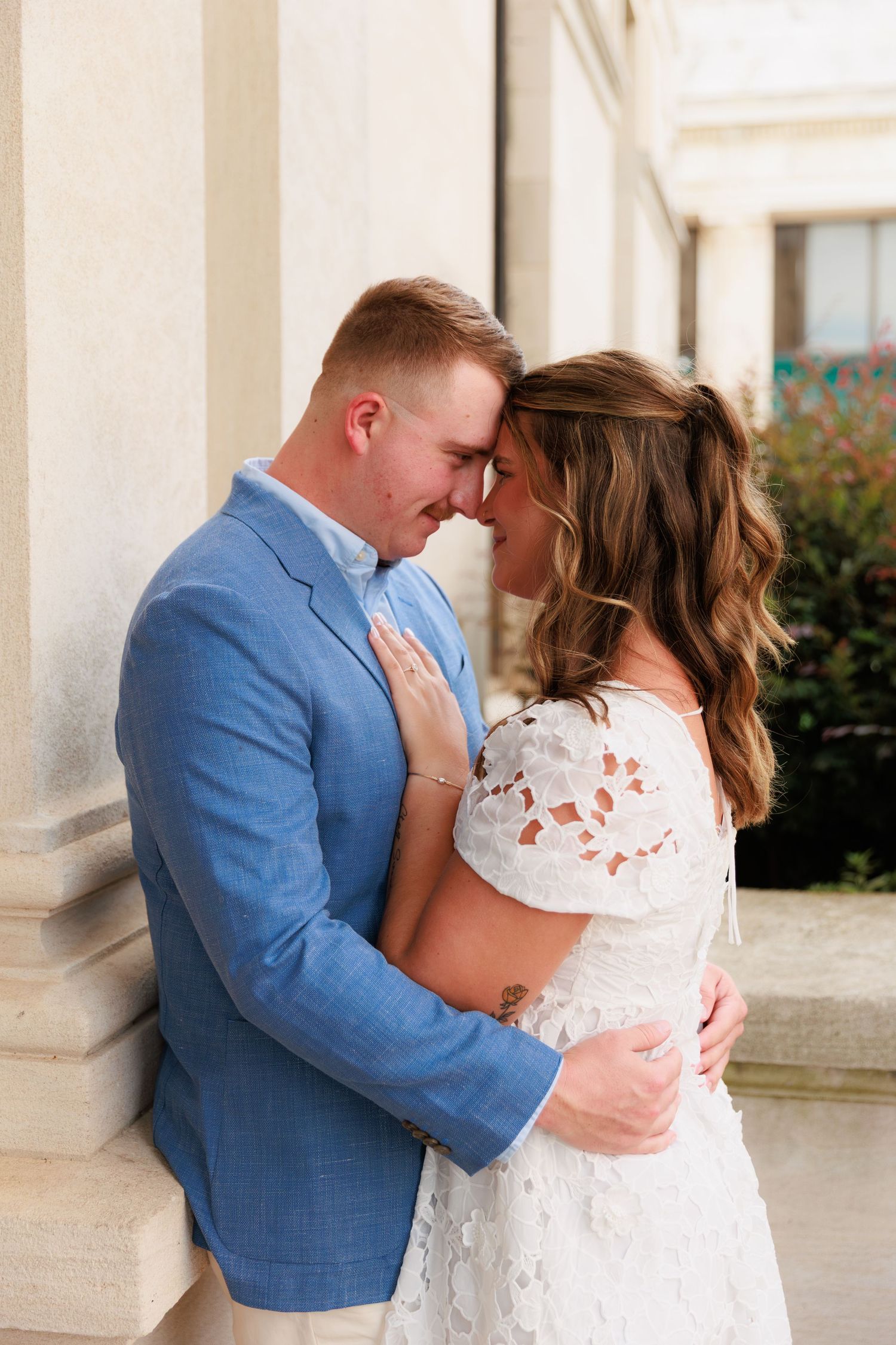 Engaged couple sharing an intimate forehead touch, man in blue blazer embracing woman in white lace dress by columns.