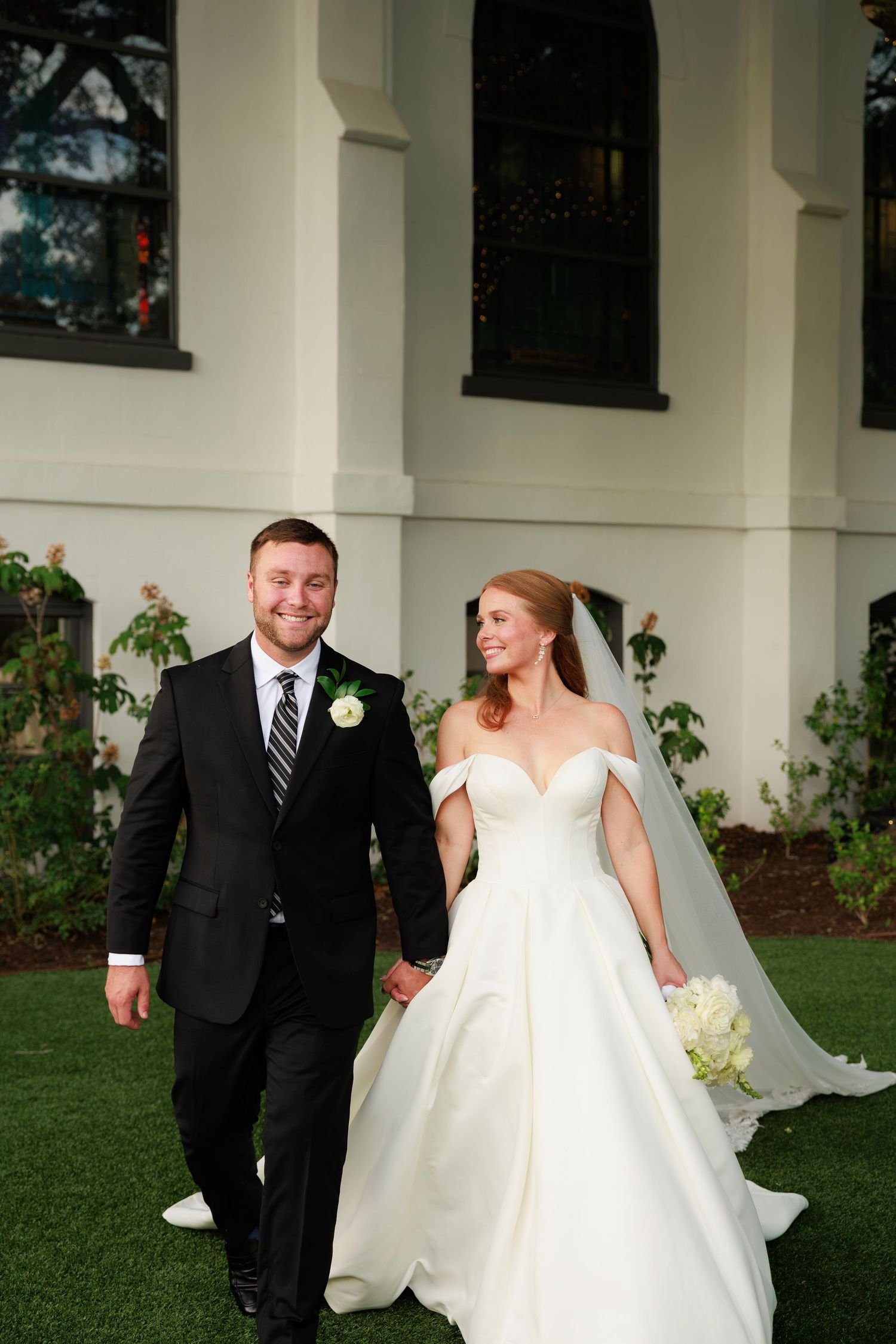 Smiling bride and groom walk hand in hand across green lawn outside elegant white wedding venue.