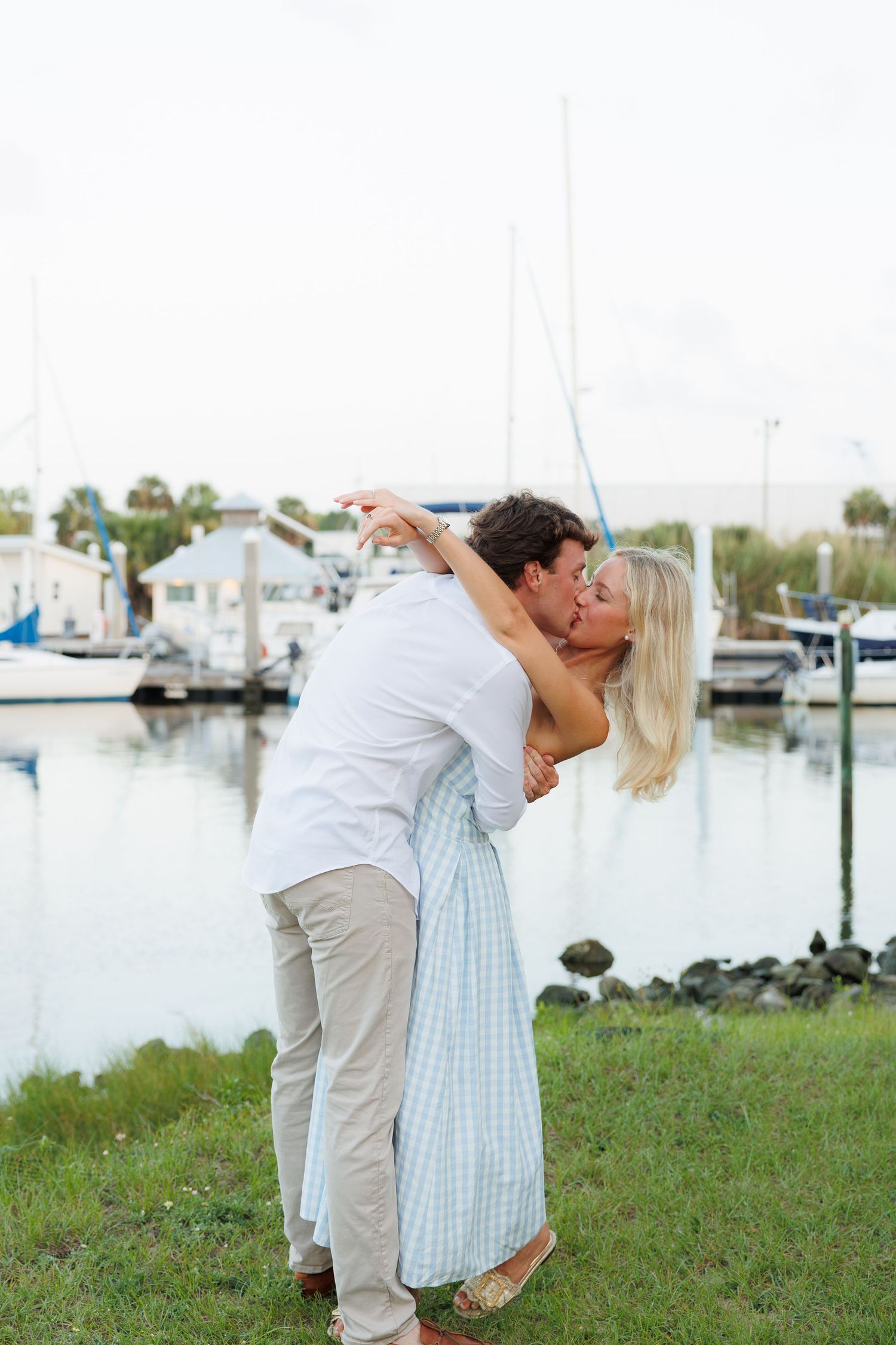 Romantic couple sharing a kiss near a marina, woman in blue gingham dress dipping back gracefully.