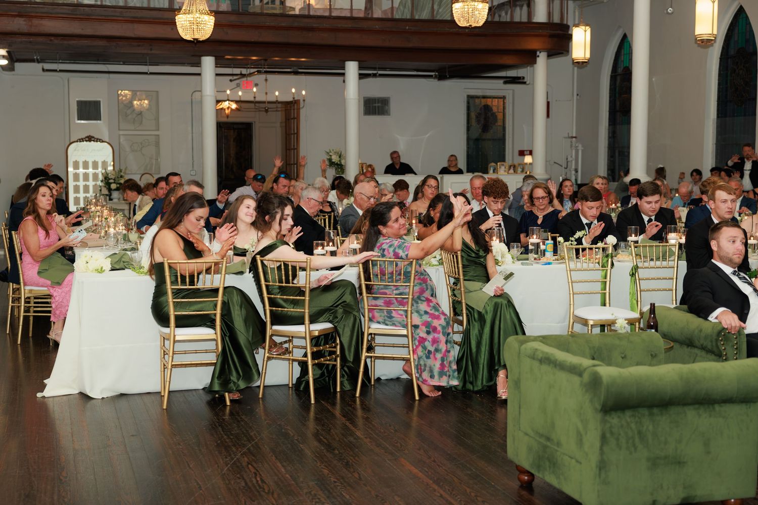 Elegant wedding reception with guests seated at green velvet chairs and white tables in a grand ballroom venue.