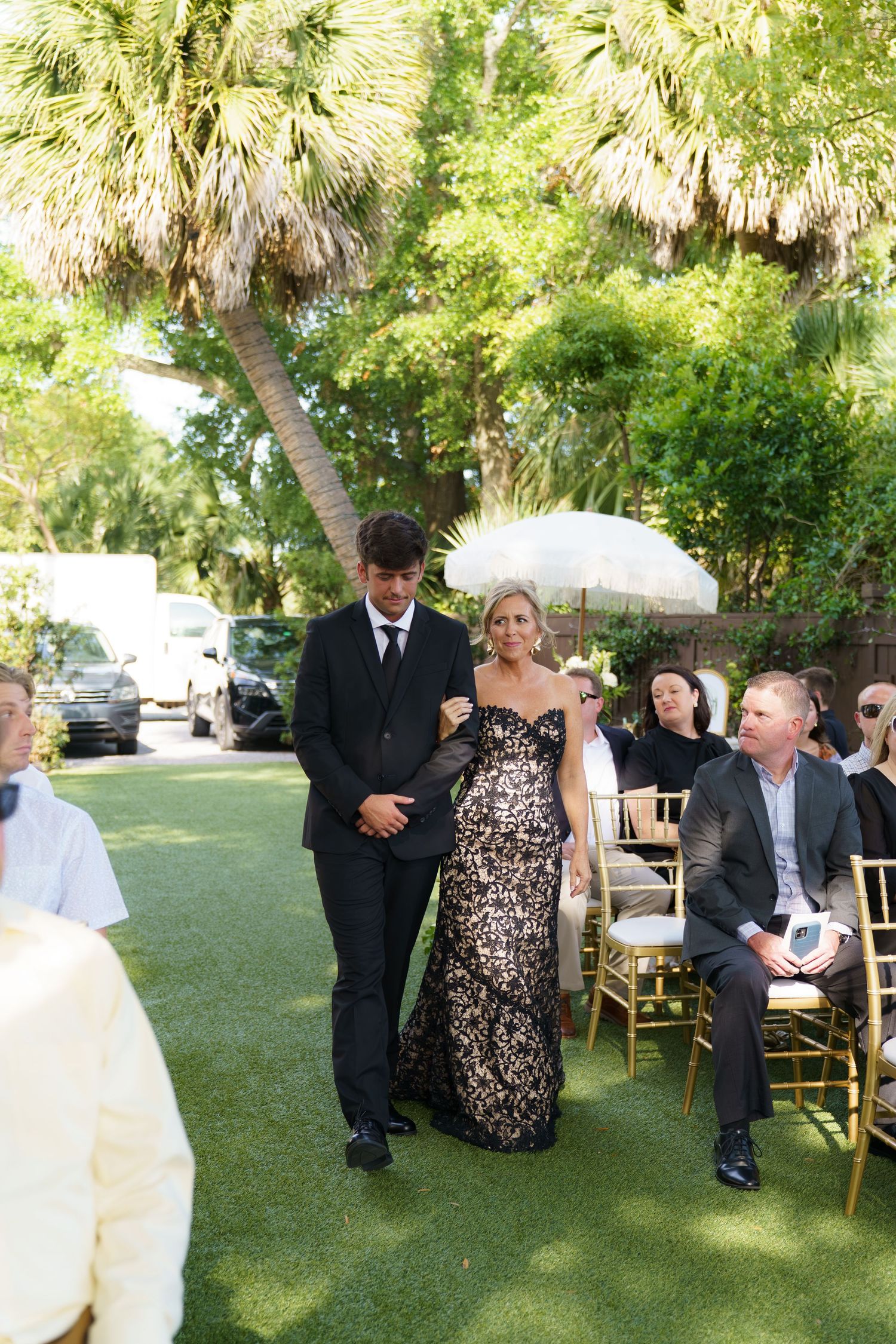 A woman in a black floral gown walks down the aisle with a man in a suit at an outdoor wedding ceremony.