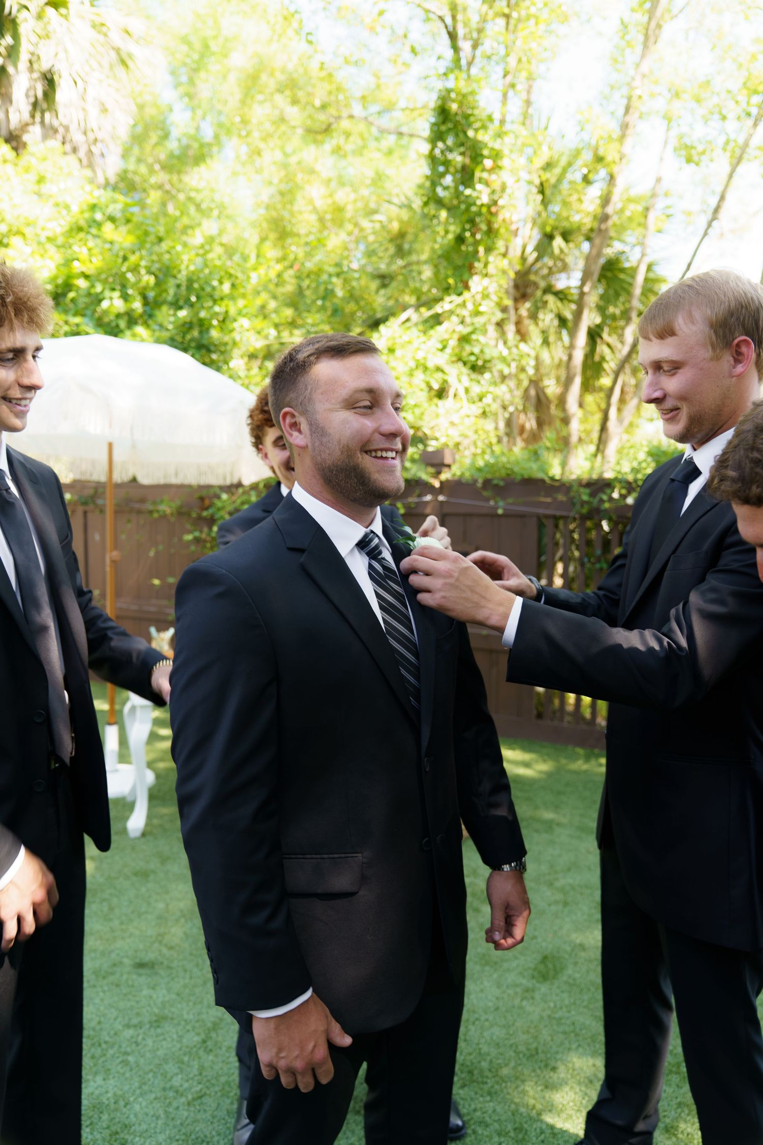 Groomsmen in black suits helping groom with his tie outdoors at a backyard wedding ceremony.