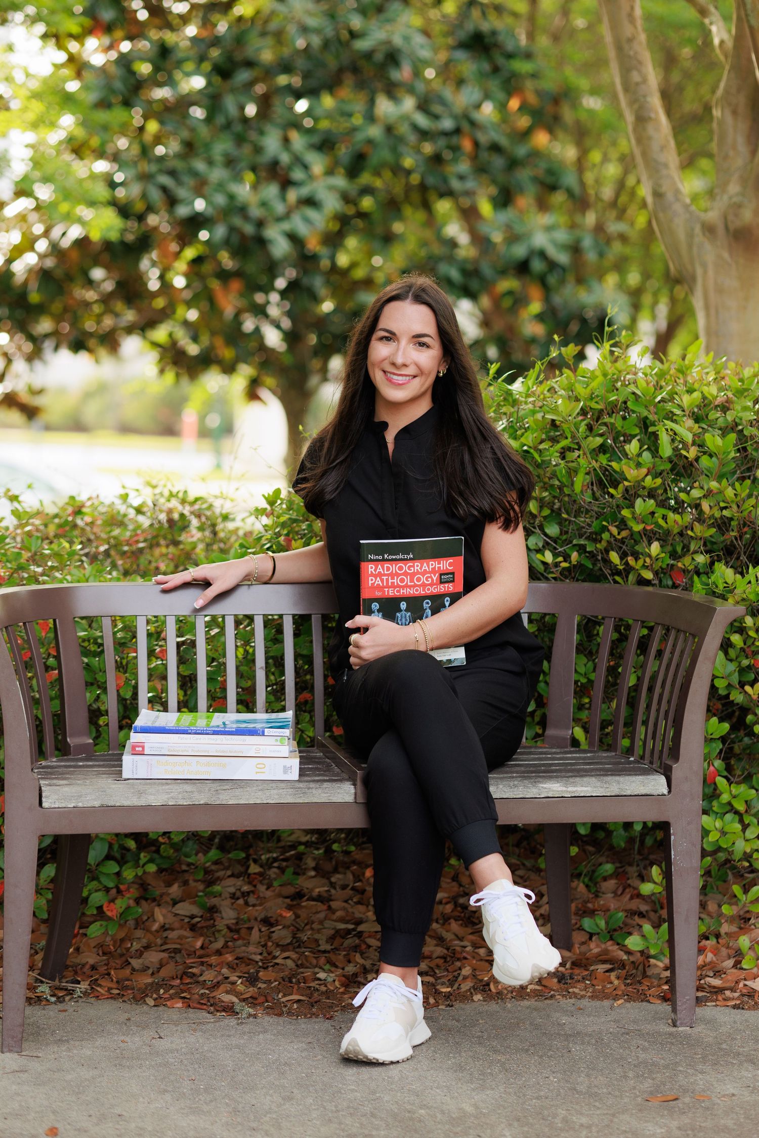 Smiling woman in black outfit sits on bench holding radiology textbook surrounded by lush green trees outdoors.