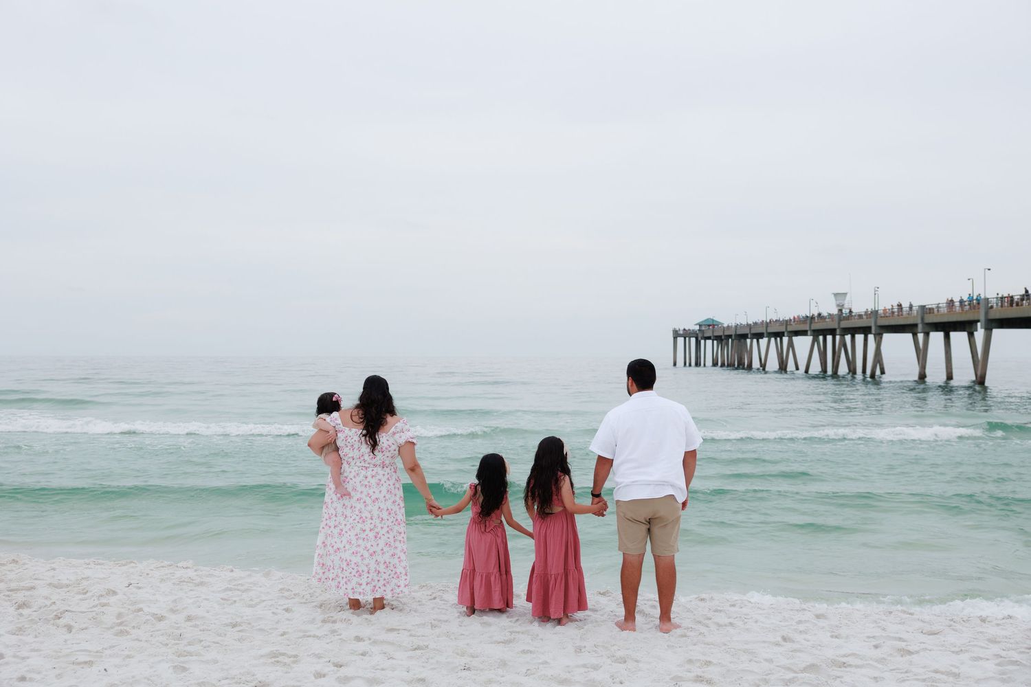 Family of four holding hands on a beach near a pier, facing the ocean on a cloudy day.