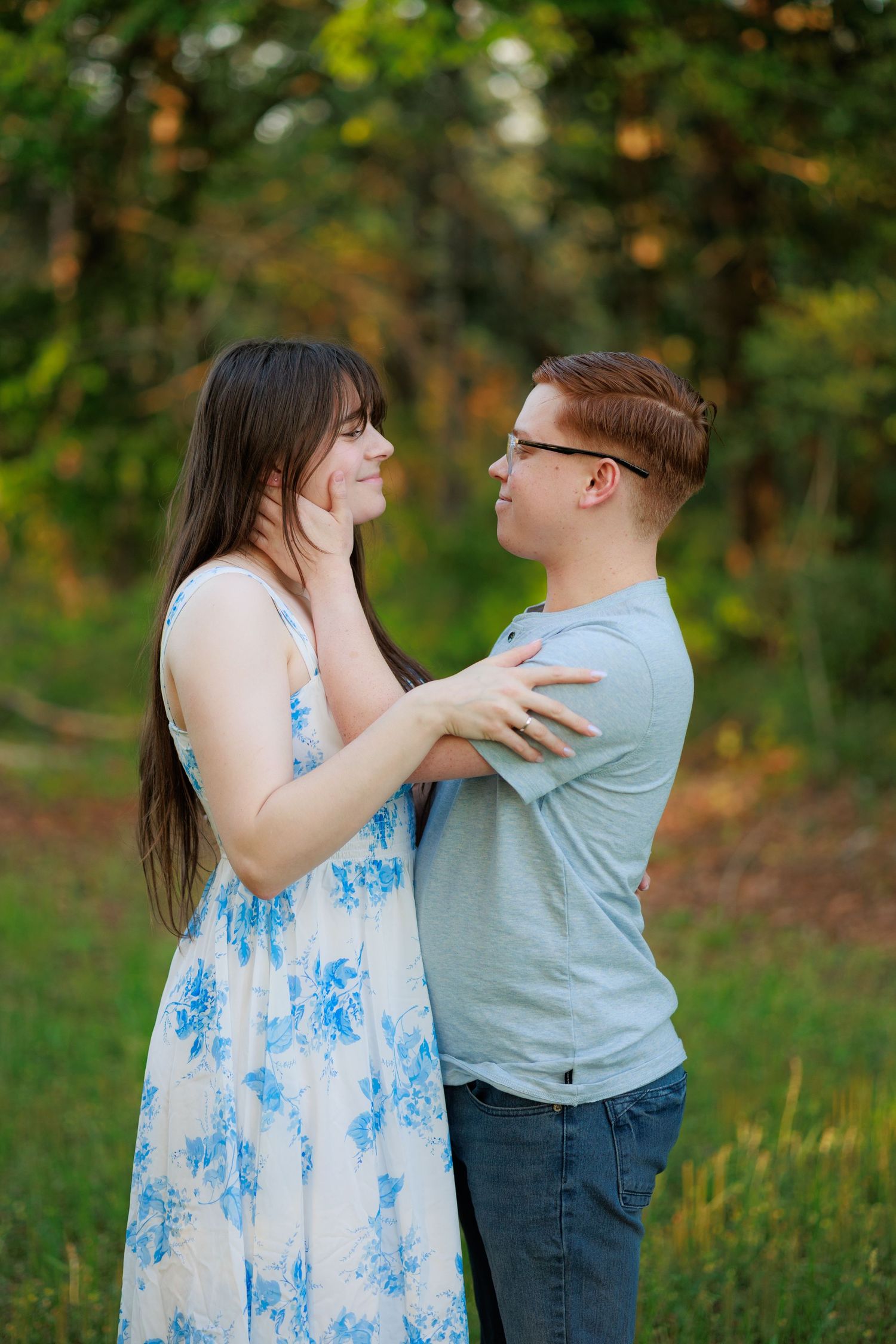 A young couple embraces and smiles at each other outdoors, she in a blue floral dress, he in a gray t-shirt.